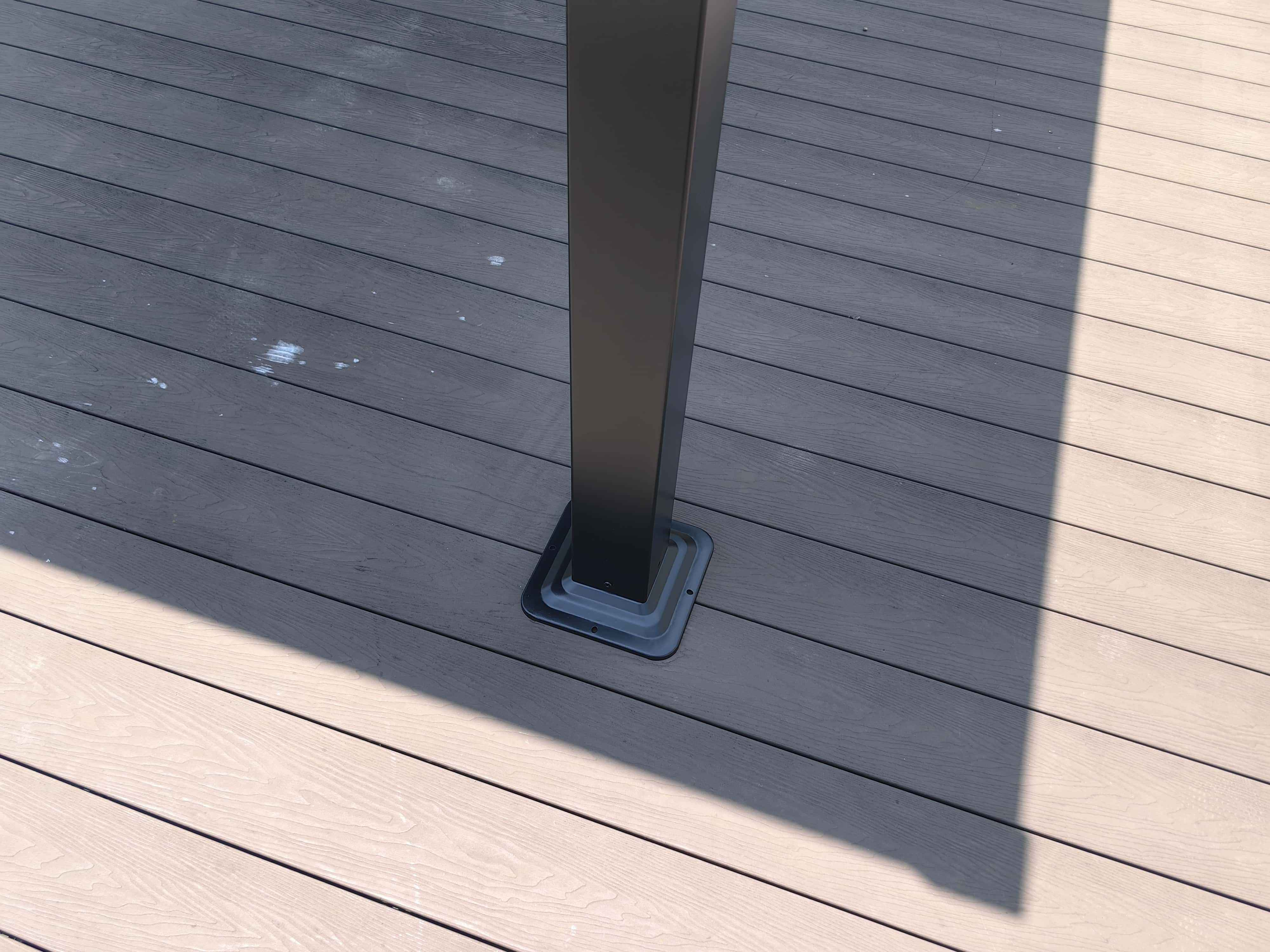 Ein dunkelgrauer Pfosten auf einem hellgrauen Holzdeck. Schatten fallen über die Bretter.