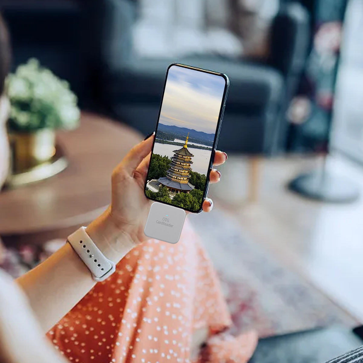 Frau, die ein Telefon hält, das eine Pagode zeigt. Eine weiße Uhr und ein Kartenleser sind ebenfalls sichtbar. Der Hintergrund ist ein Wohnzimmer.