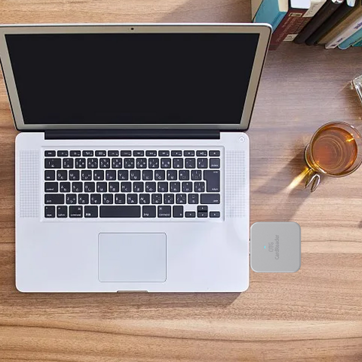Ein Laptop mit schwarzem Bildschirm steht auf einem Holztisch. Ein Kartenleser und ein Glas Tee daneben, Bücher im Hintergrund.
