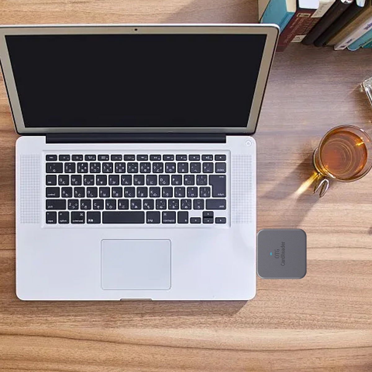 Laptop mit Tastatur auf einem Holztisch. Eine Tasse Tee und ein Kartenleser liegen daneben. Bücher sind im Hintergrund.