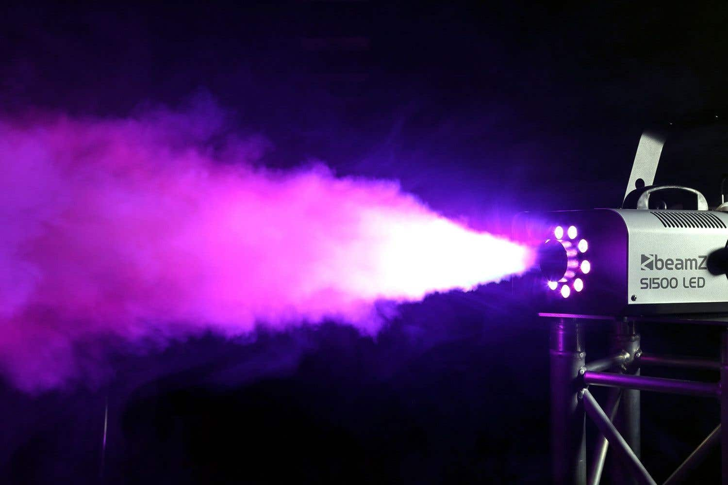 Una máquina de niebla Beamz S1500 LED emite humo púrpura. El humo está iluminado con luces brillantes sobre un fondo oscuro.