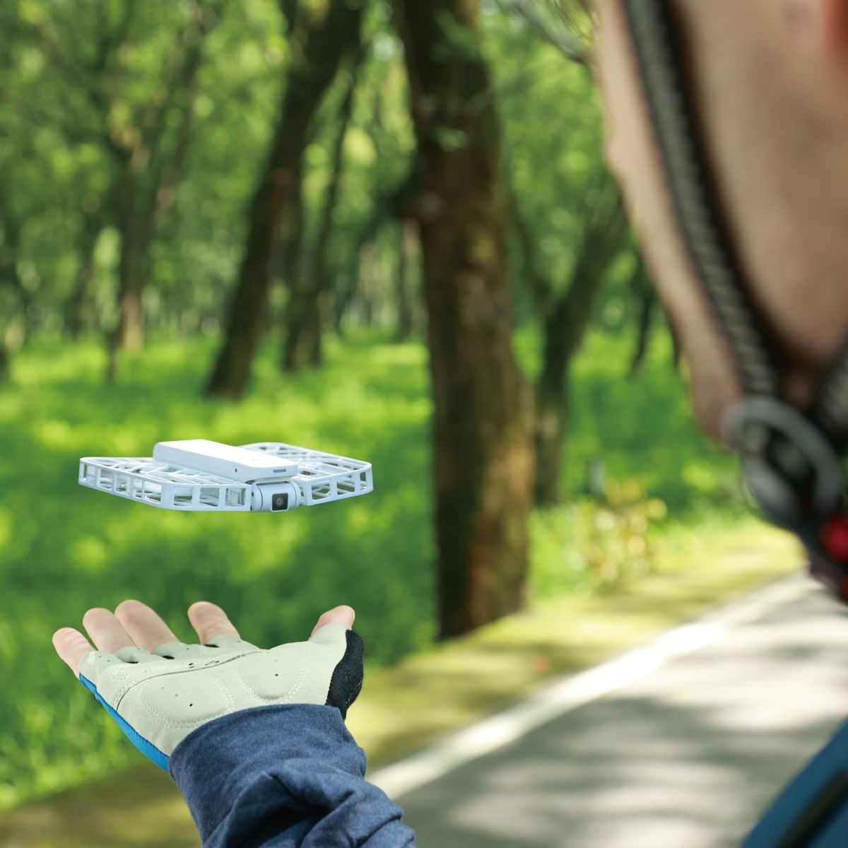 Una persona con casco sostiene un dron en un parque. El dron es blanco, y el fondo son árboles verdes y hierba.