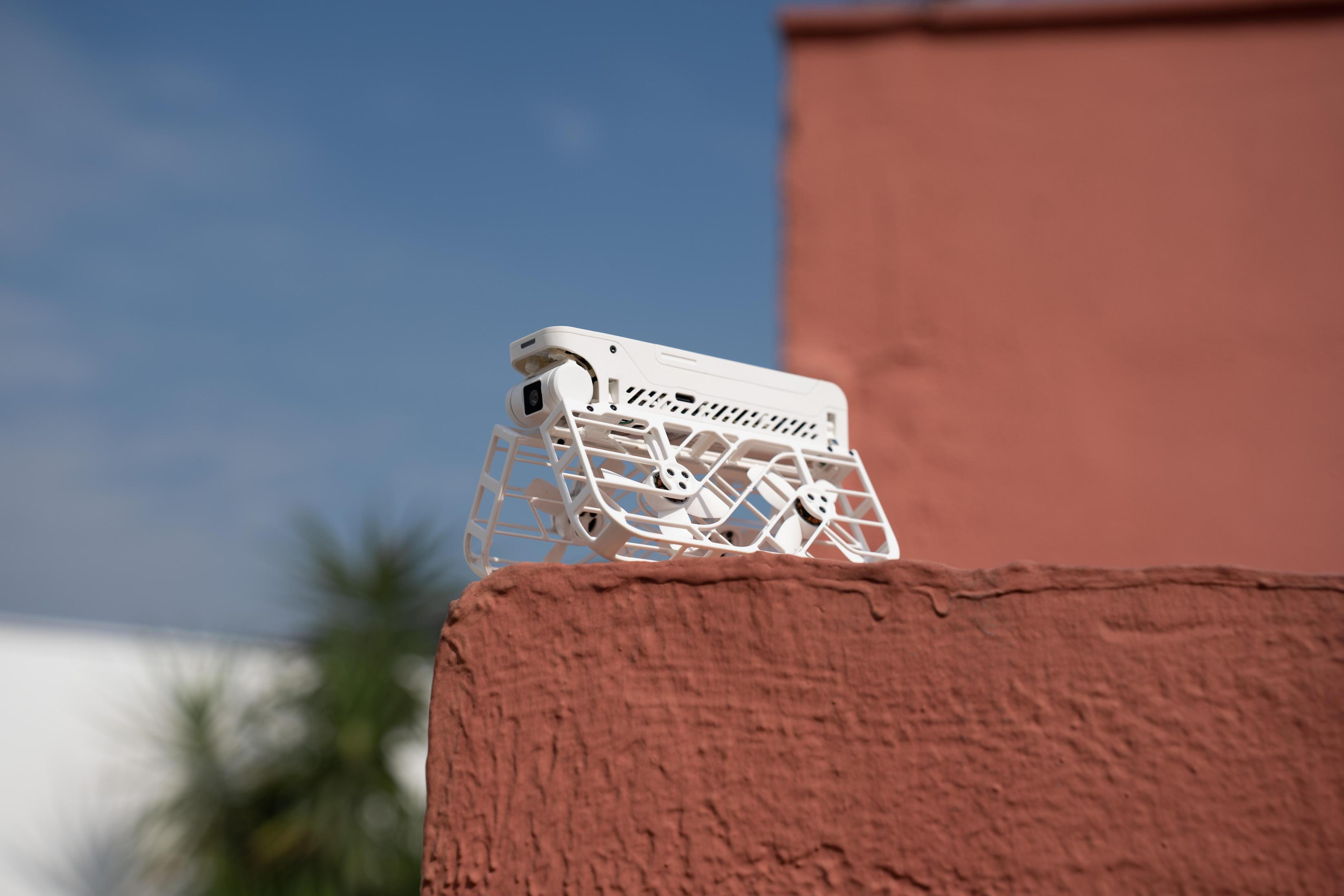 Un dron blanco con jaula protectora descansa sobre una superficie roja texturizada, contra un cielo azul y una pared roja.
