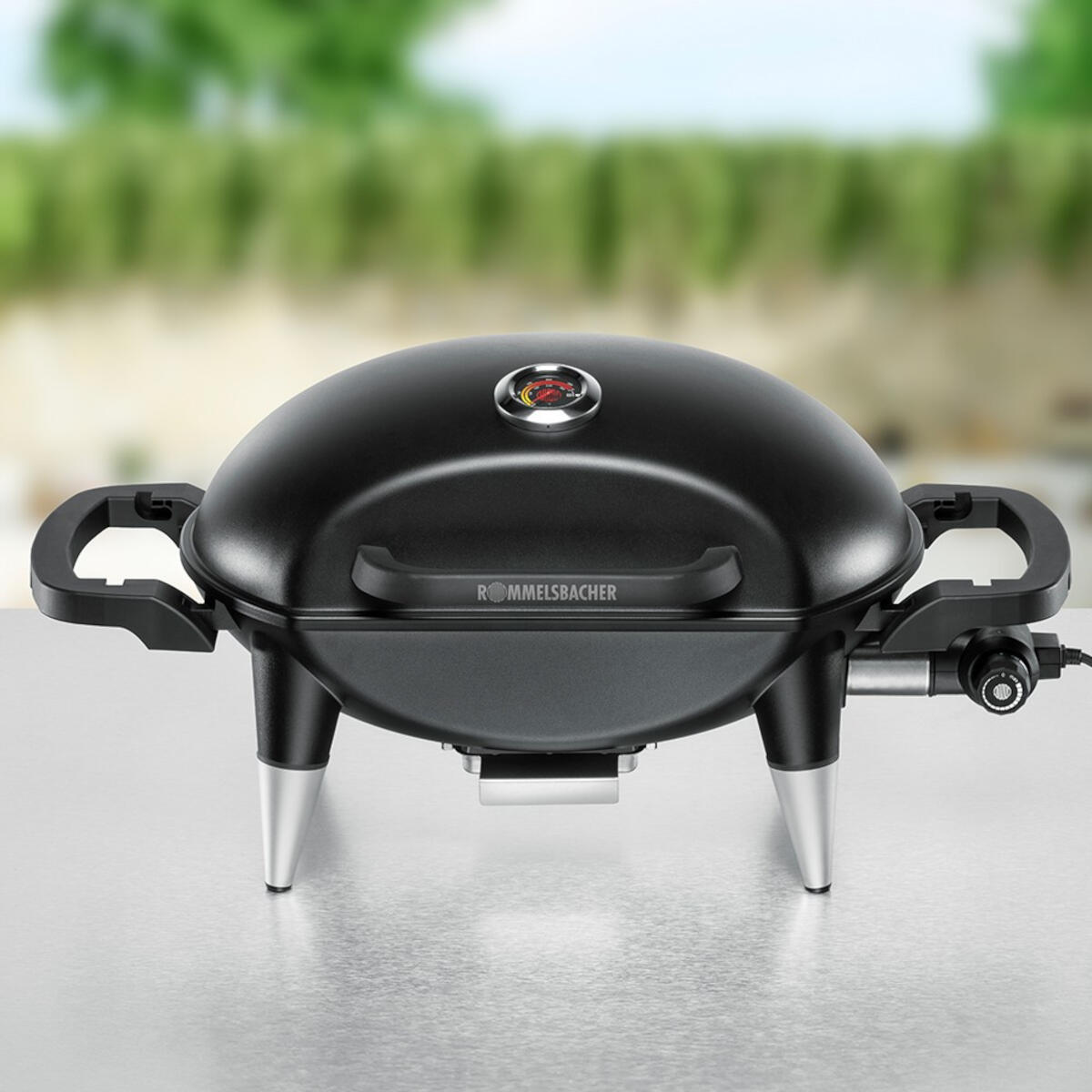 A black grill with silver legs on a metal surface. The background is blurred green foliage.