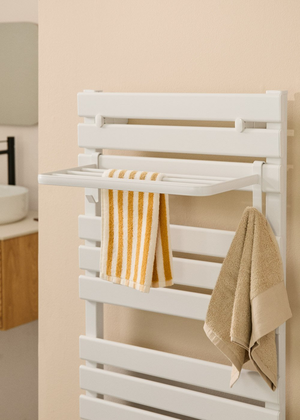 Toallero blanco con dos toallas. Una toalla es a rayas amarillas y blancas, la otra beige. El fondo es una pared beige claro.