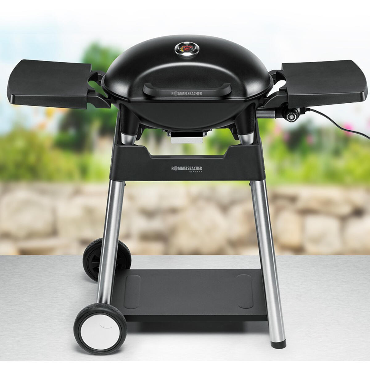 Un grill nero RÖMMSLBACHER su un supporto con ruote. Ha tavoli laterali, un coperchio e un ripiano inferiore. Lo sfondo è sfocato.