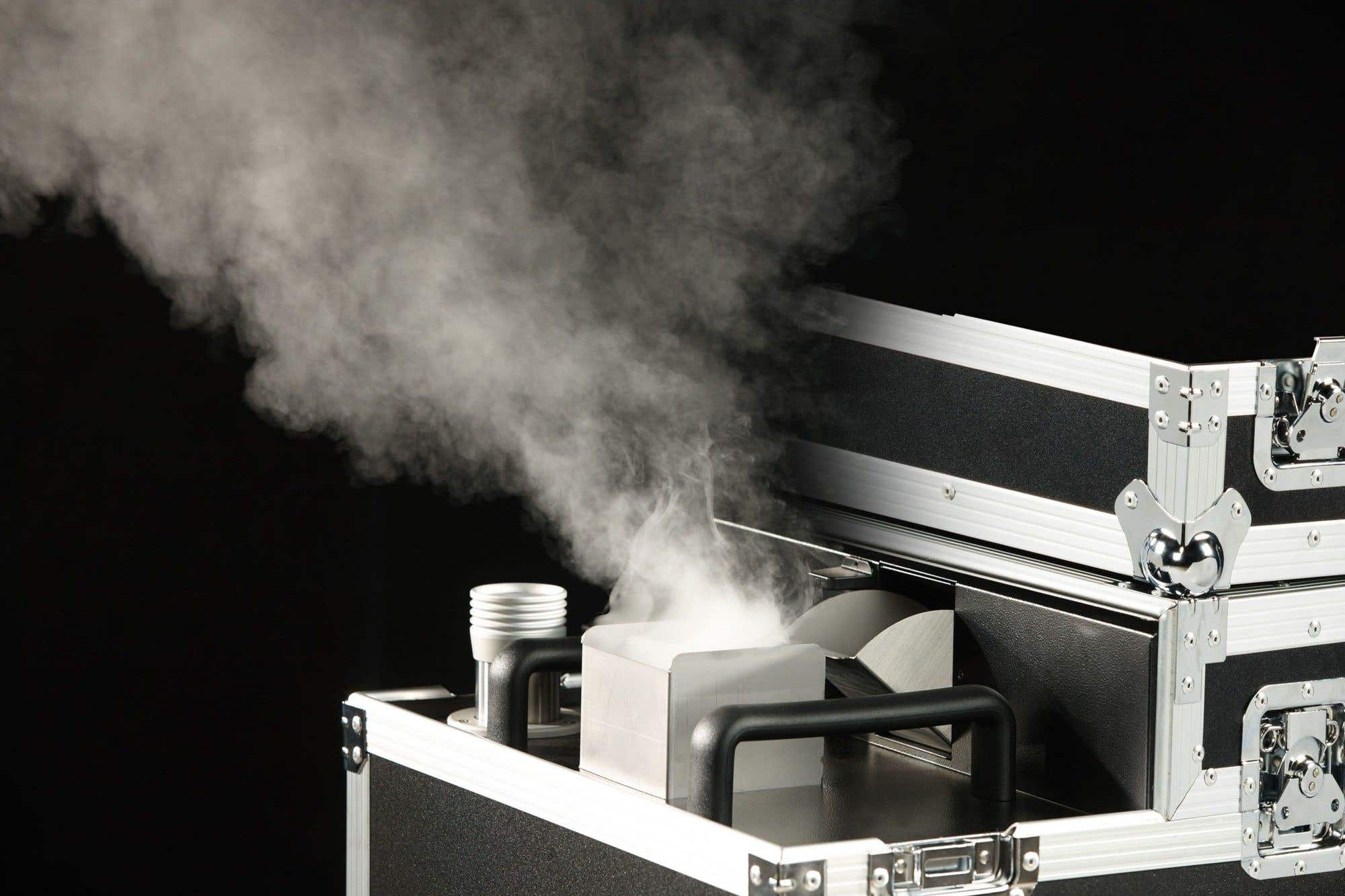 Una máquina de humo emite humo blanco. La máquina está dentro de una caja negra con bordes metálicos, sobre un fondo negro.