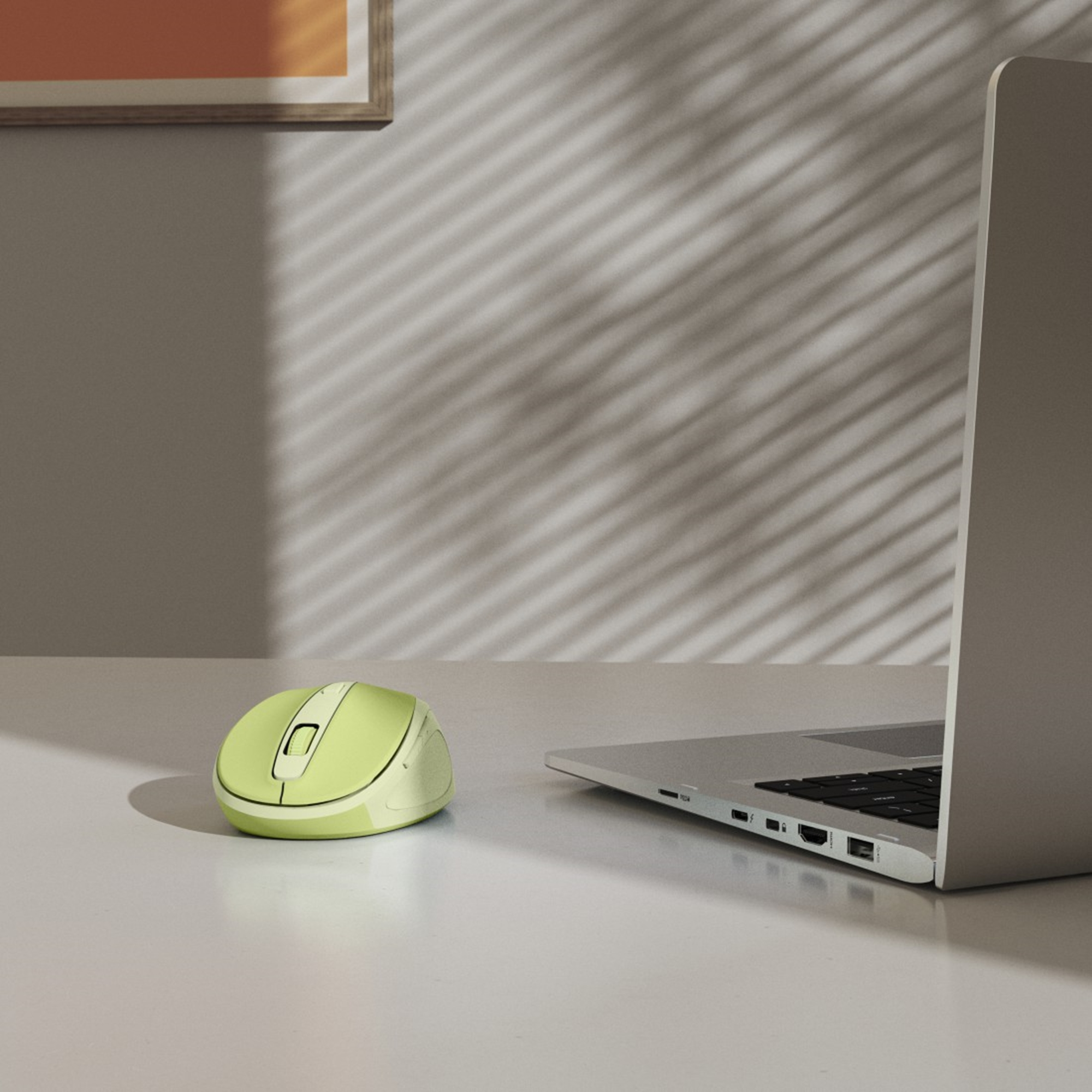 Un mouse wireless verde lime accanto a un laptop argentato su una scrivania bianca. Le ombre delle persiane sono sul muro.