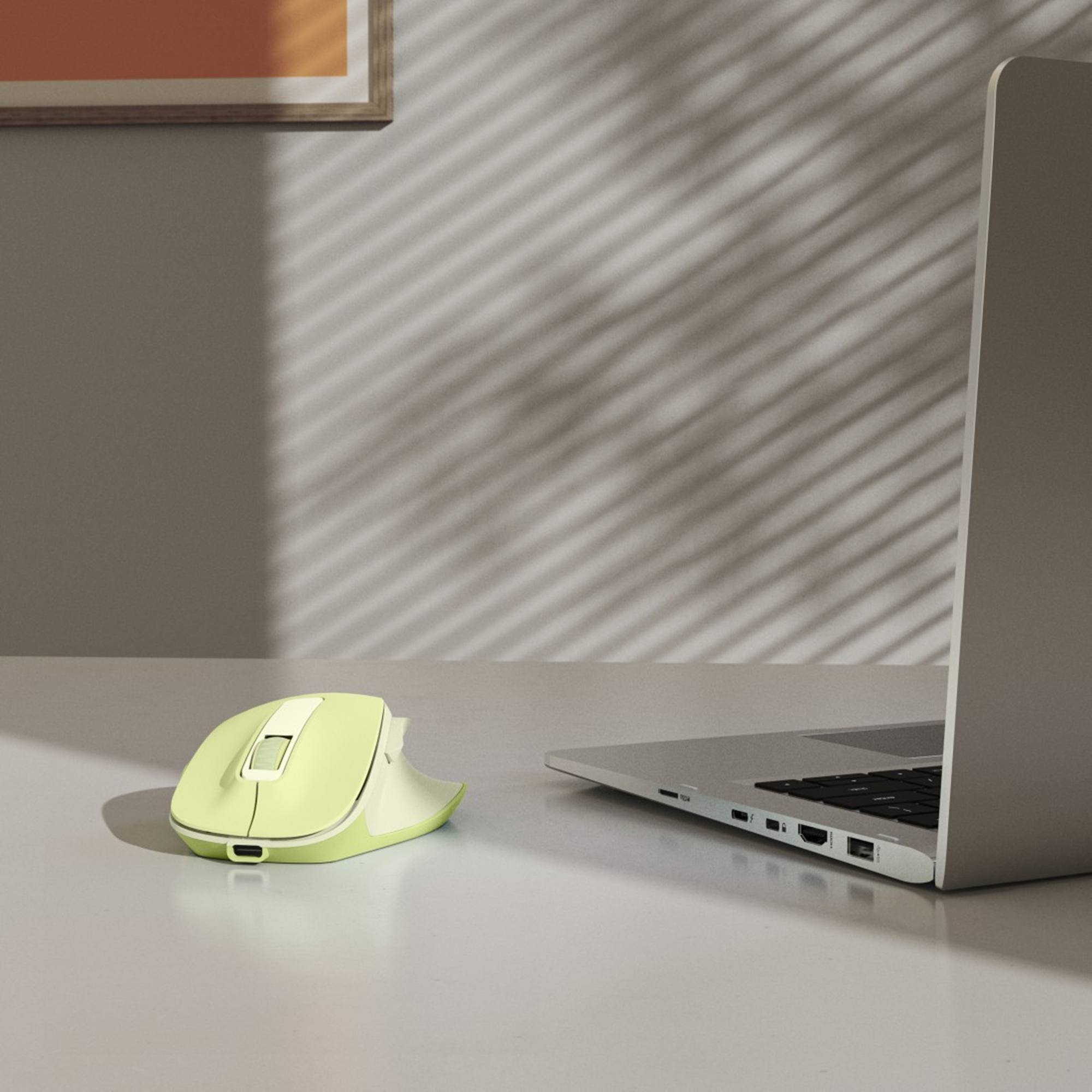Eine gelb-weiße kabellose Maus liegt neben einem silbernen Laptop auf einer weißen Oberfläche. Schatten sind auf der Wand.