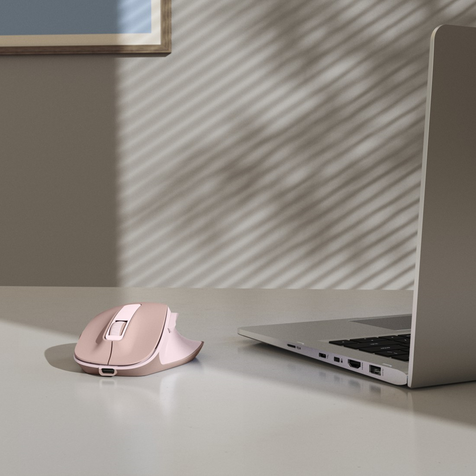 Eine roségoldene Maus liegt neben einem Laptop auf einer weißen Oberfläche. Schatten vom Fenster sind an der Wand.