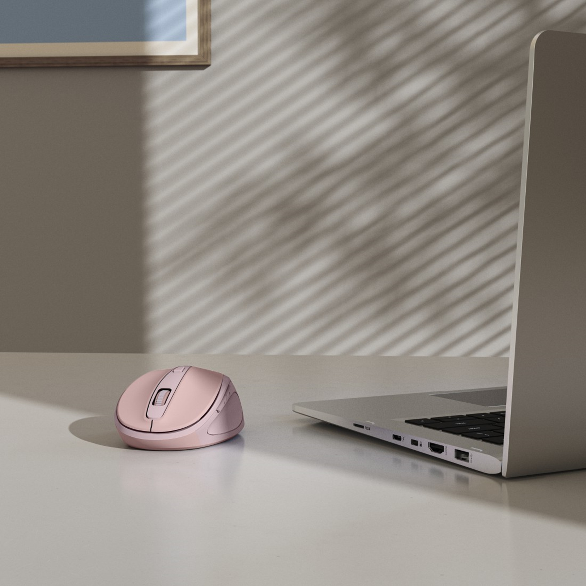 Un mouse per computer rosa è vicino a un laptop su una superficie bianca. Le ombre di una finestra sono sul muro.
