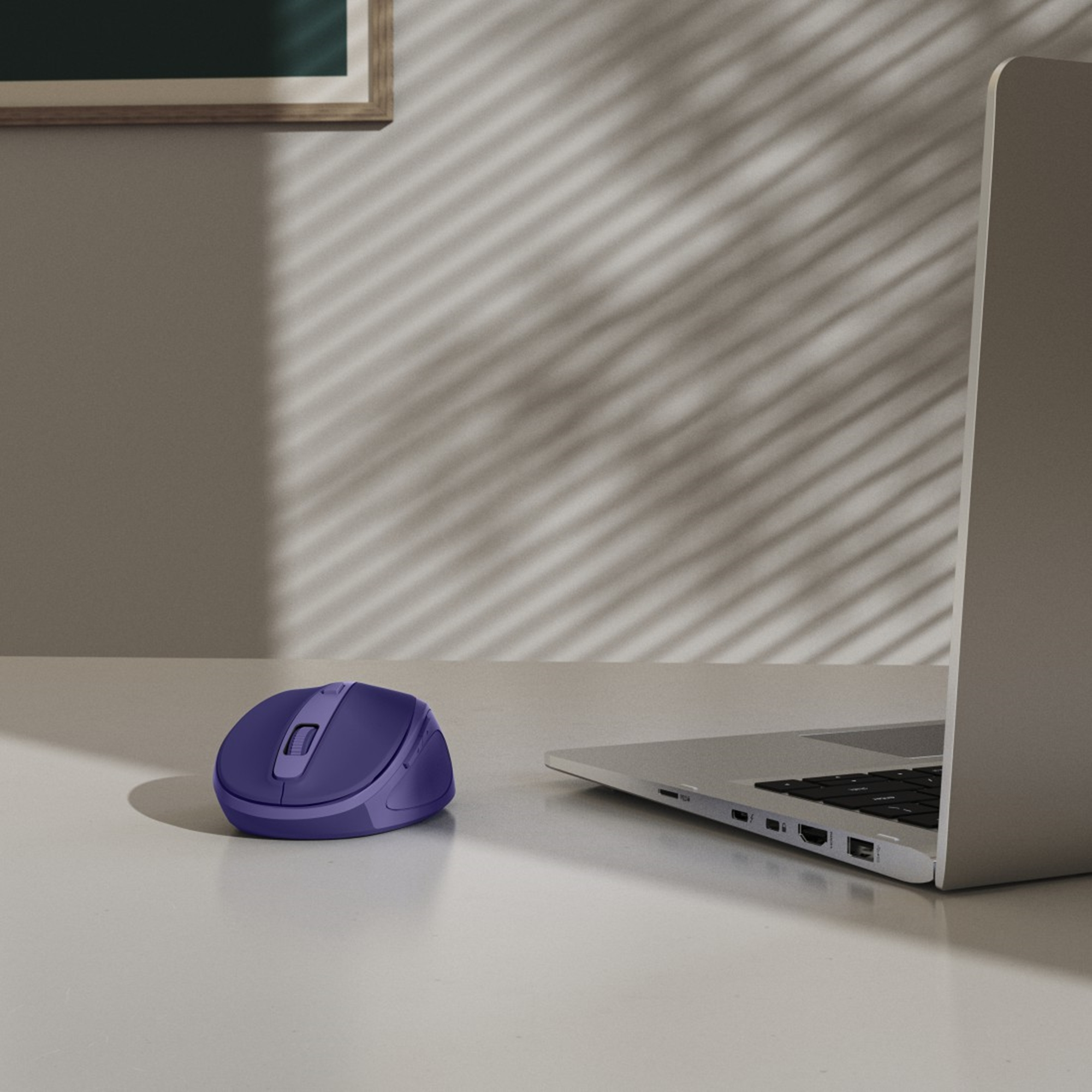 Un mouse per computer viola è appoggiato su una scrivania bianca accanto a un laptop. Le ombre cadono sul muro sullo sfondo.