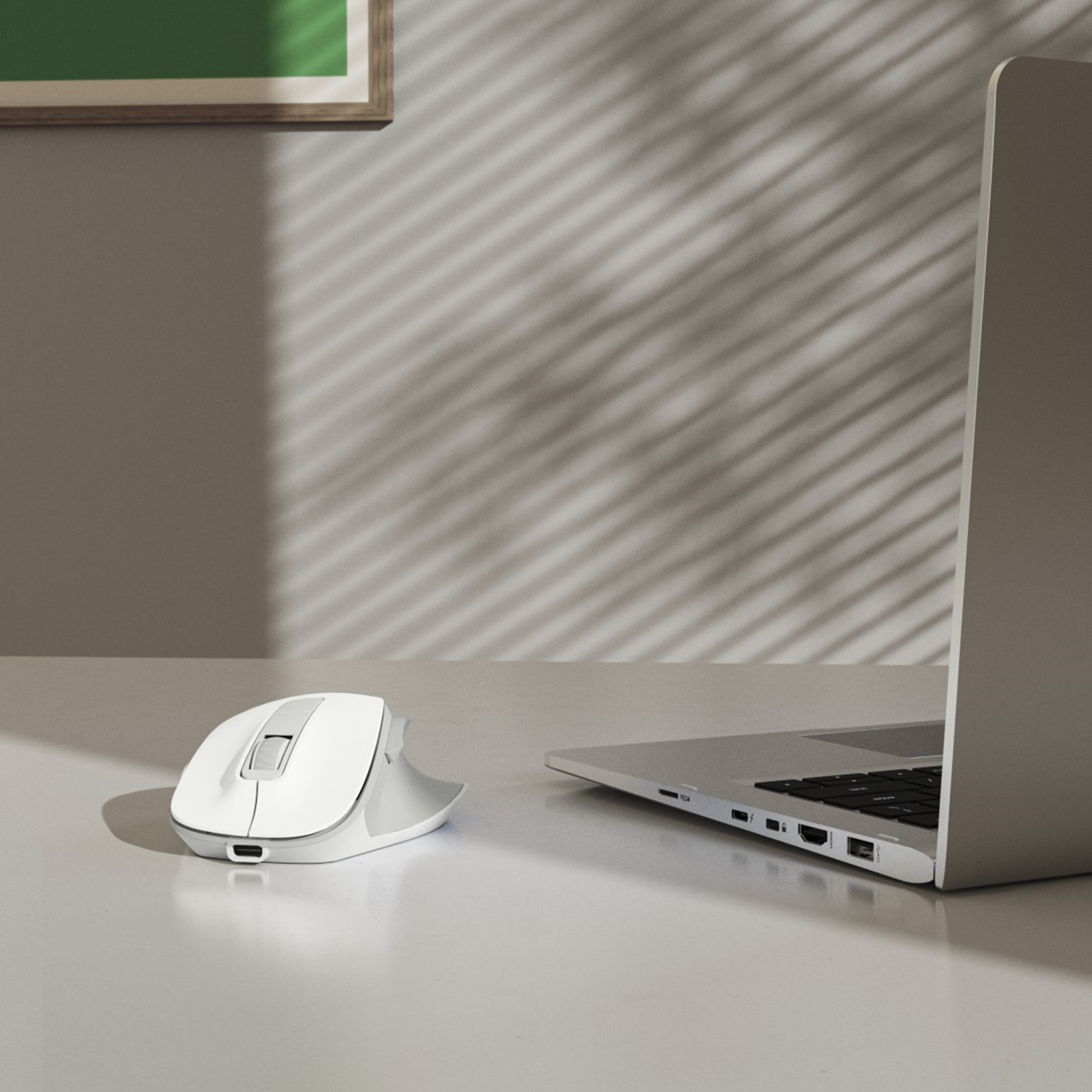 Eine weiße Maus neben einem offenen Laptop auf einer weißen Oberfläche. Schatten von Jalousien an der Wand.