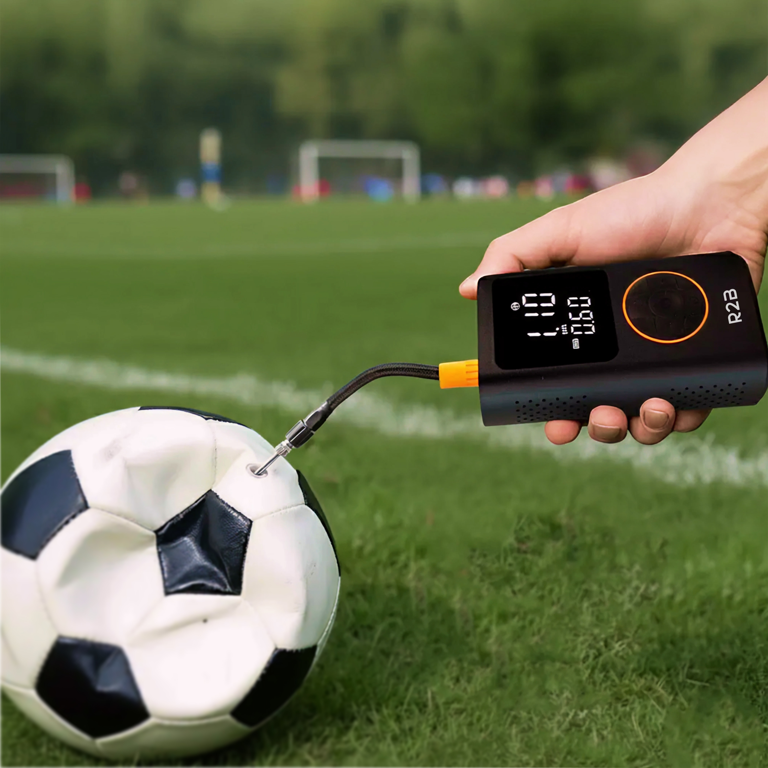 A hand inflates a soccer ball with a black and orange pump on a green field. The pump displays pressure readings.
