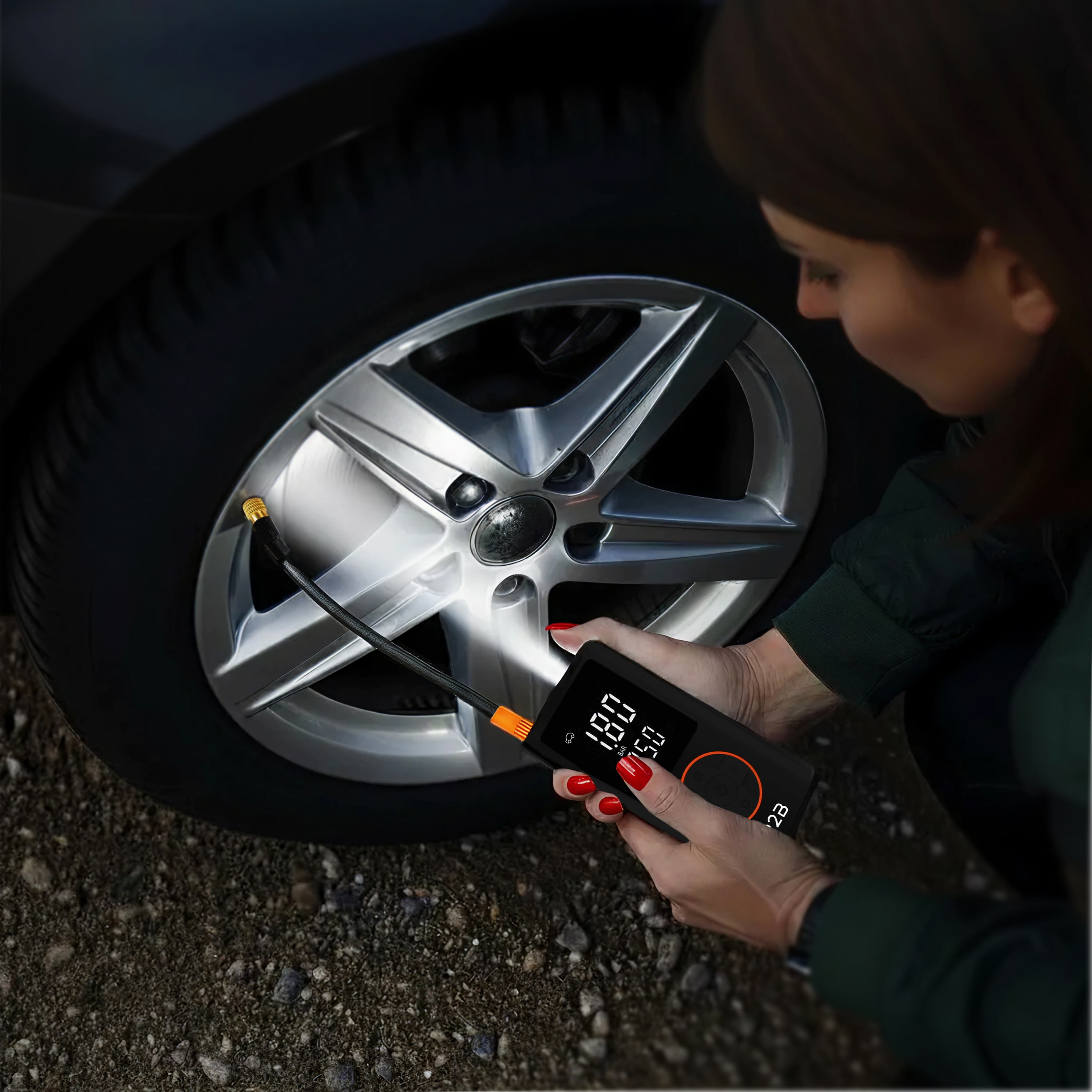 A woman inflates a tire with a digital gauge. The tire is mounted on a silver alloy wheel. Asphalt is on the ground.