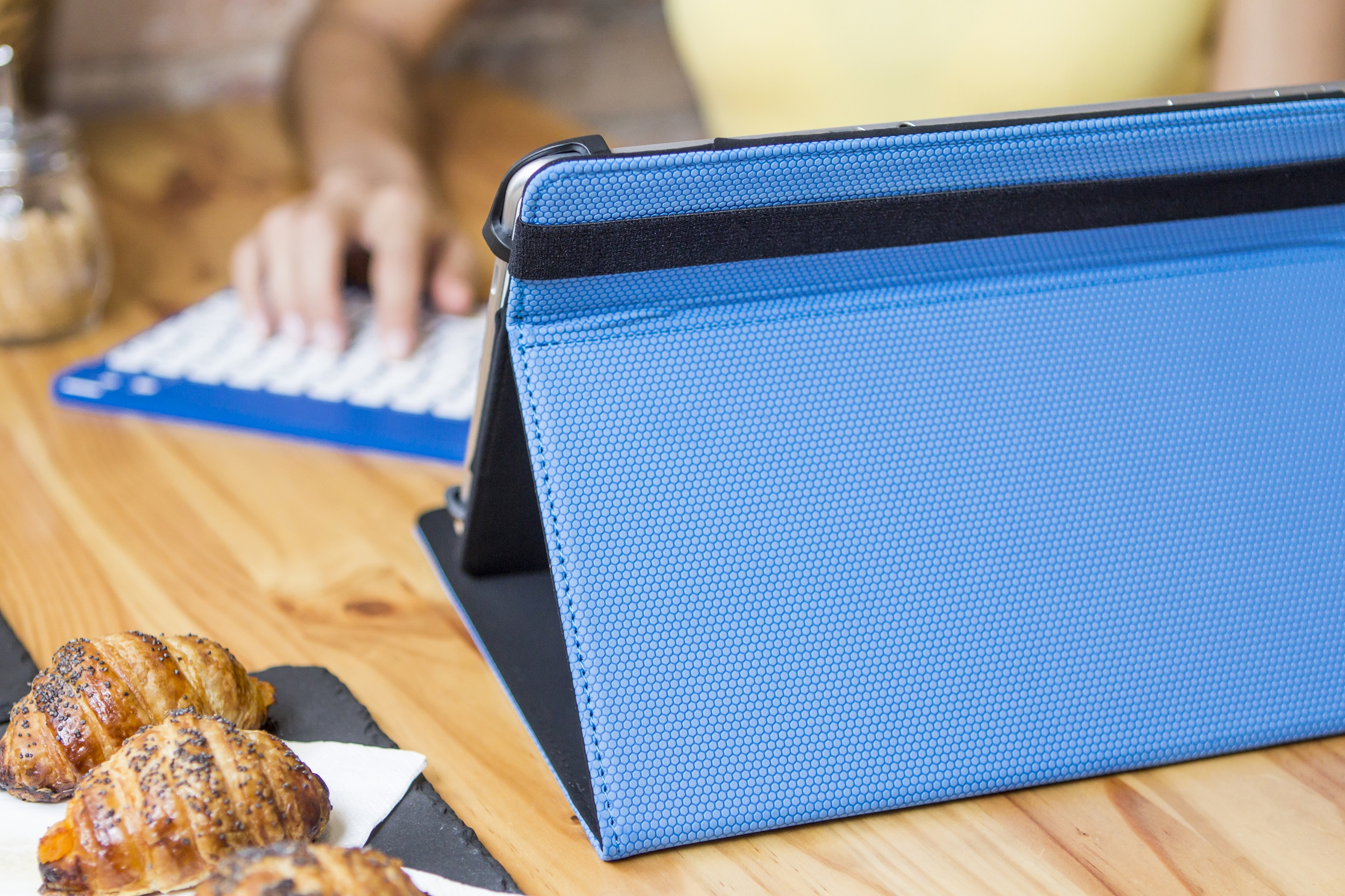 Funda azul para tableta, cruasanes y teclado sobre mesa de madera. Una persona teclea.