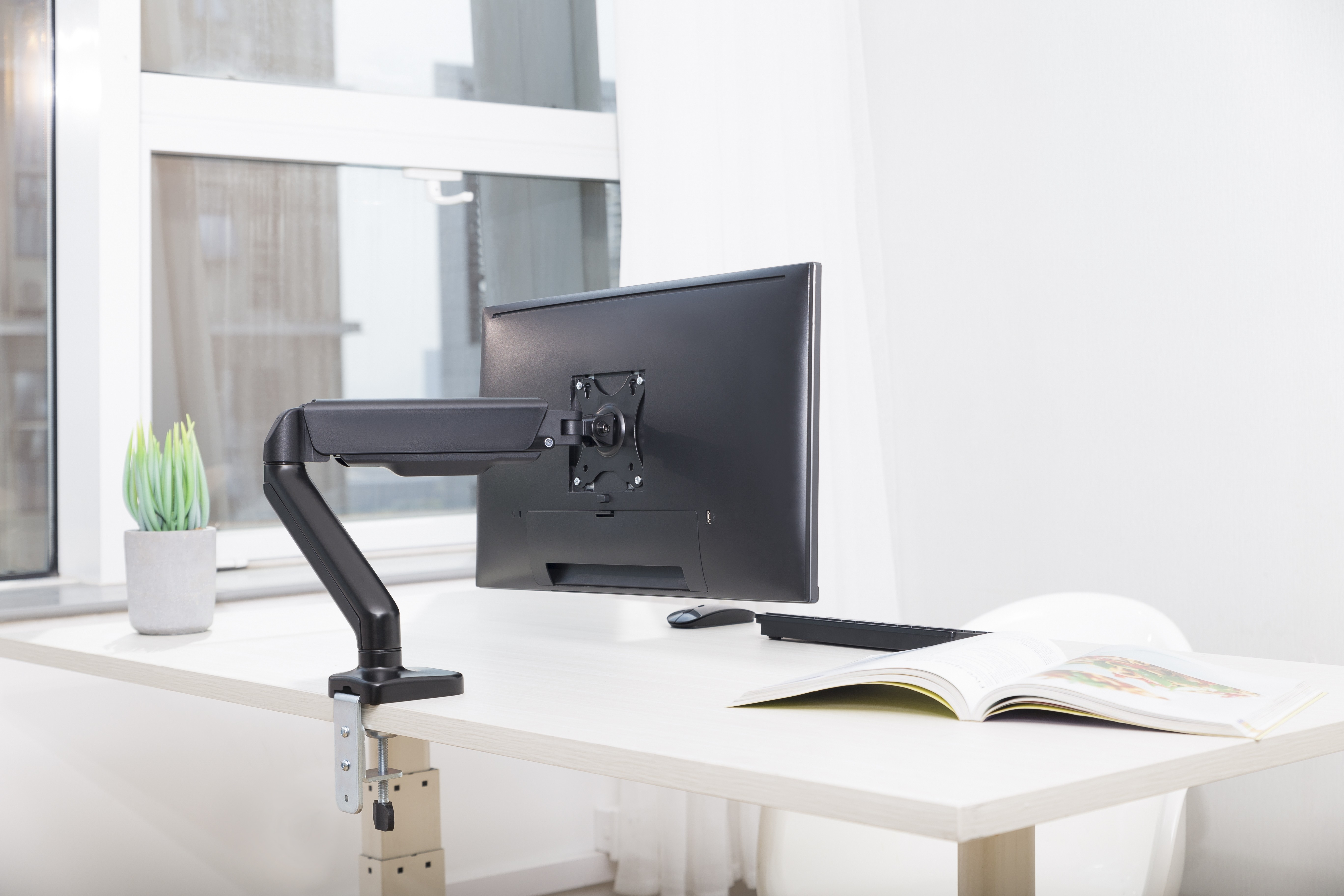 A computer monitor on a desk, with a book and a plant. The monitor is black and mounted on an arm.