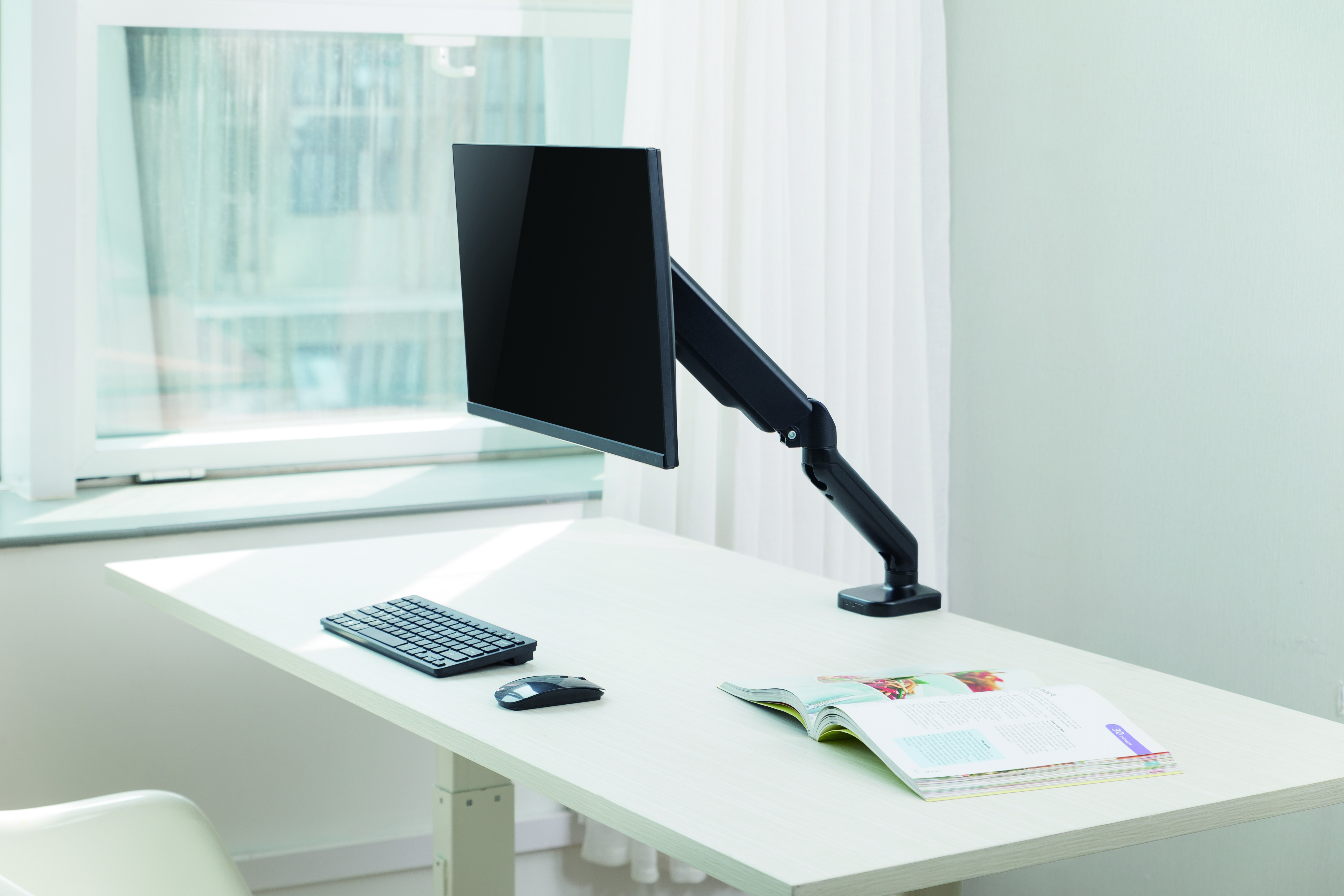 A desk with a monitor, keyboard, mouse, and an open book. A window and white curtain are in the background.