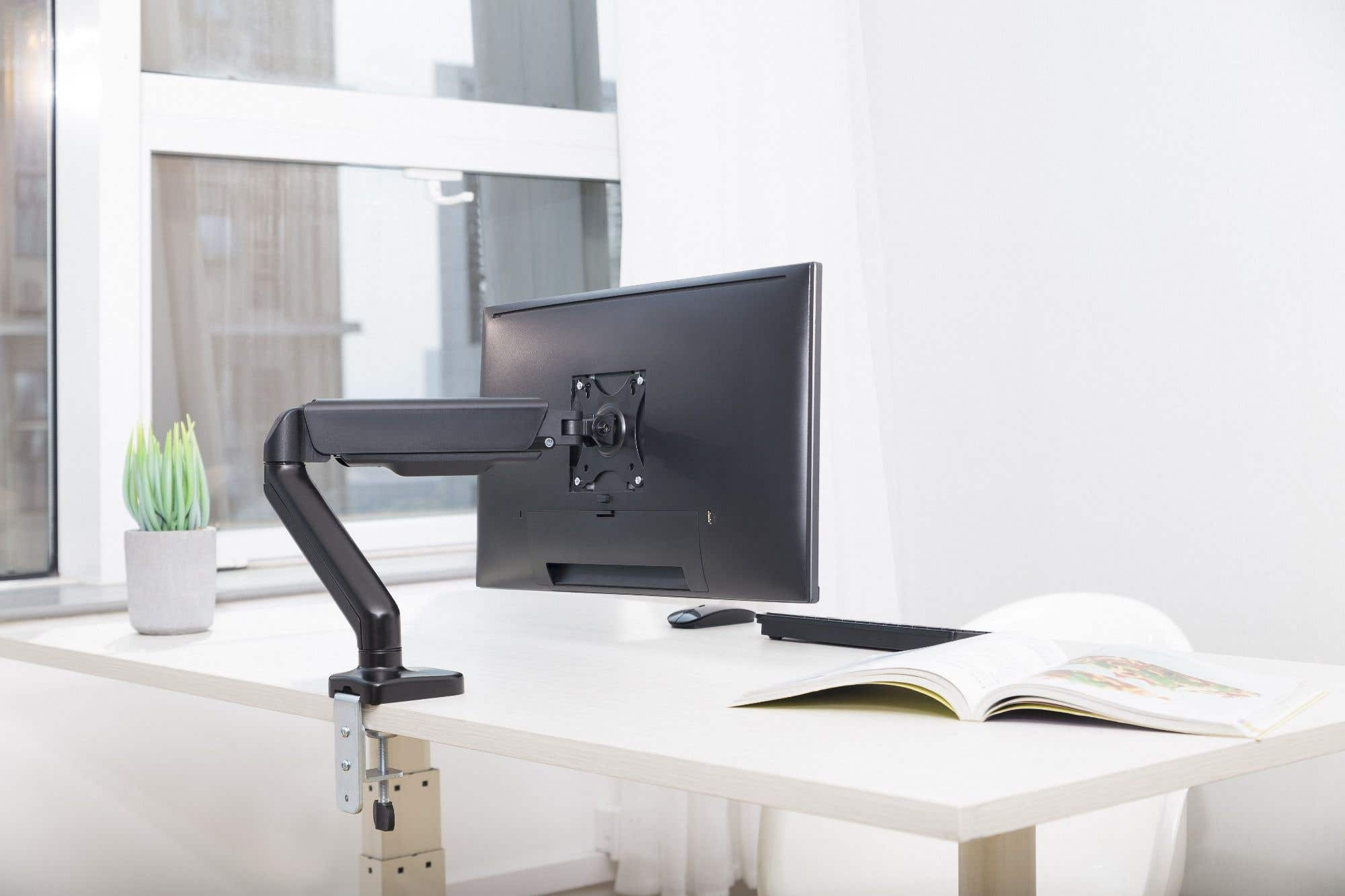 A computer monitor mounted on a desk. A book and a mouse are on the desk. A plant is on the left.