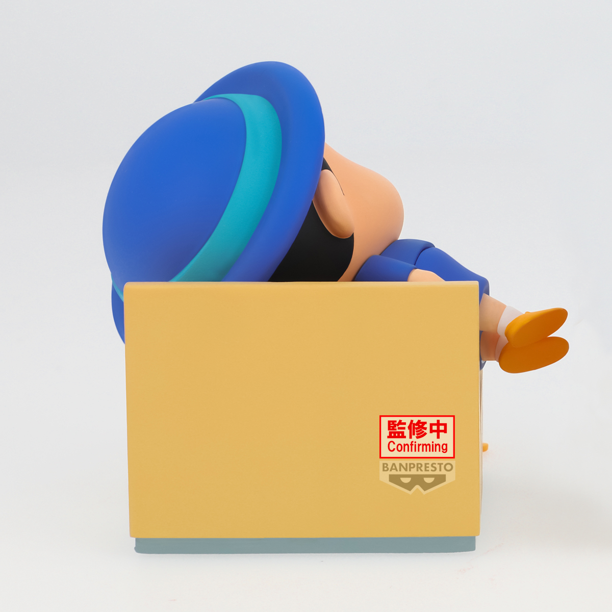Un personaggio dei cartoni animati con un cappello blu, appoggiato su una scatola gialla. La scatola ha testo giapponese e un logo Banpresto.
