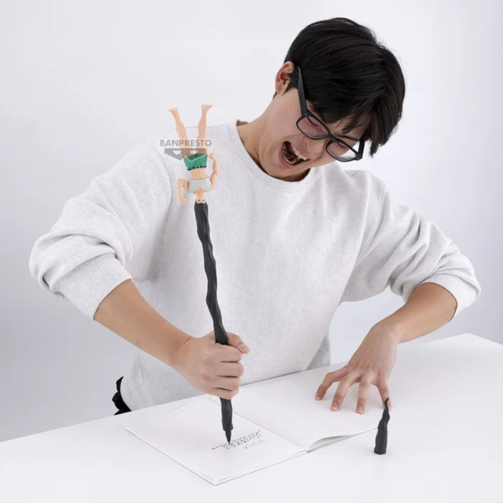 Ein Mann mit Brille schreibt mit einem Stift, der eine Figur hat. Er lächelt. Ein Notizbuch und eine kleine Figur liegen auf dem Tisch.