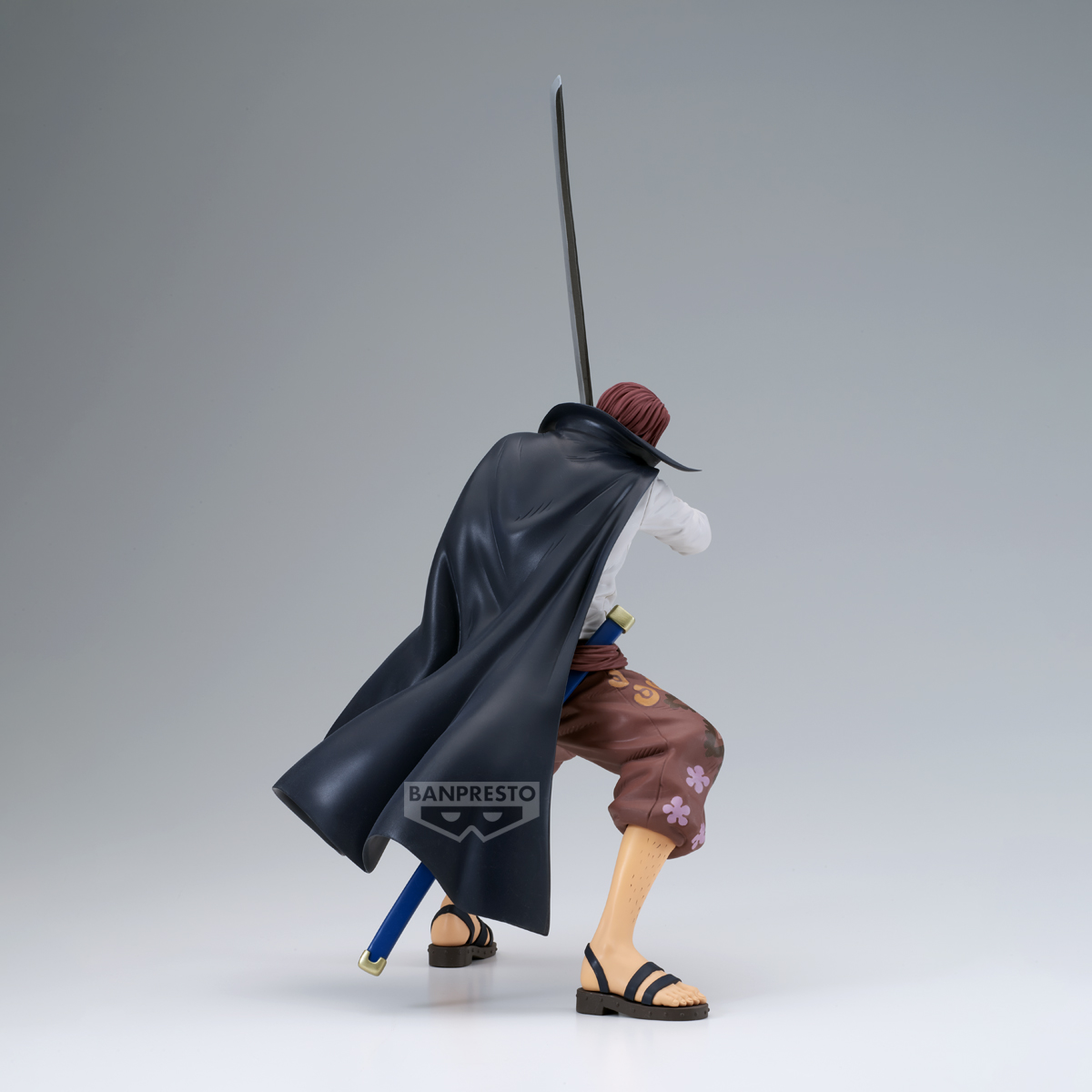 Figura de hombre con pelo rojo, capa negra y espada, de espaldas. Viste pantalón marrón, sandalias y camisa blanca.