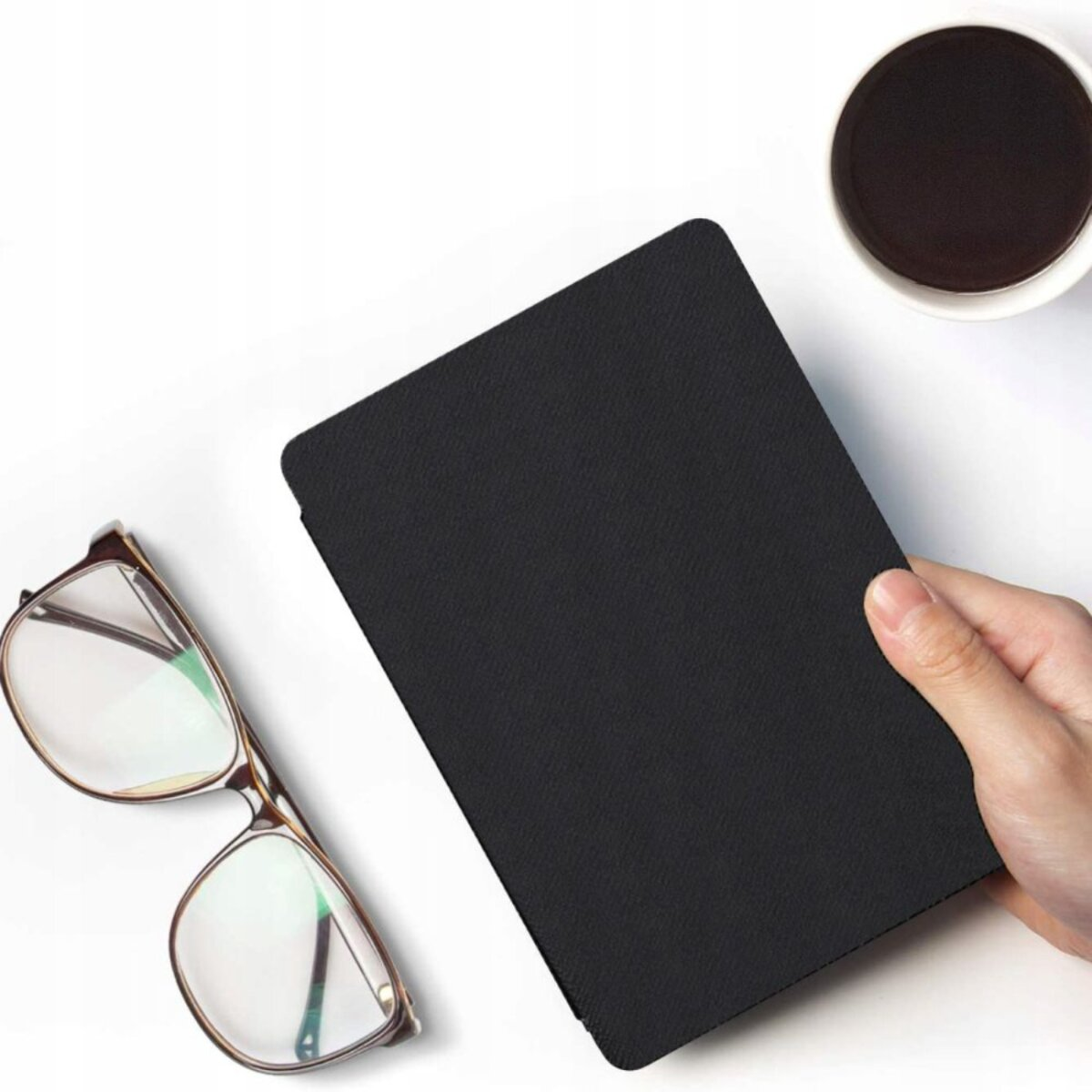 Eine Hand hält ein schwarzes Tablet. Brille und eine Tasse Kaffee liegen auf weißem Untergrund.