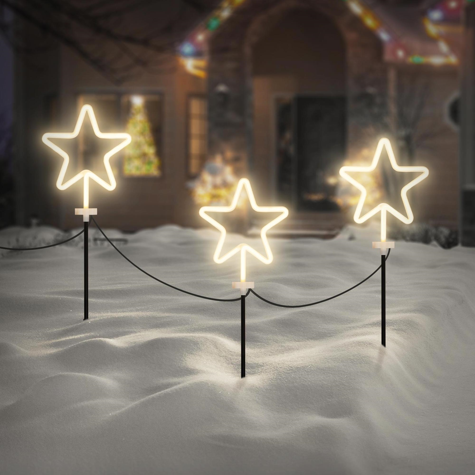 Drei beleuchtete Sternverzierungen im Schnee. Im Hintergrund ein Haus mit Lichtern.