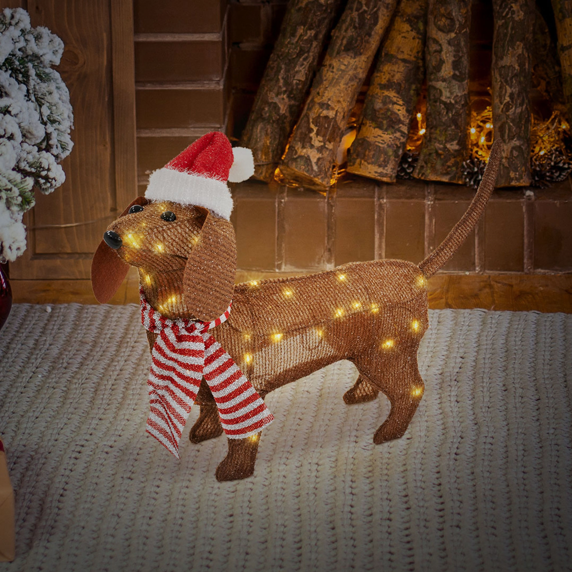 Ein dekorativer Dackel mit Weihnachtsmütze und Schal, beleuchtet mit Lichtern, steht auf einer Strickdecke.