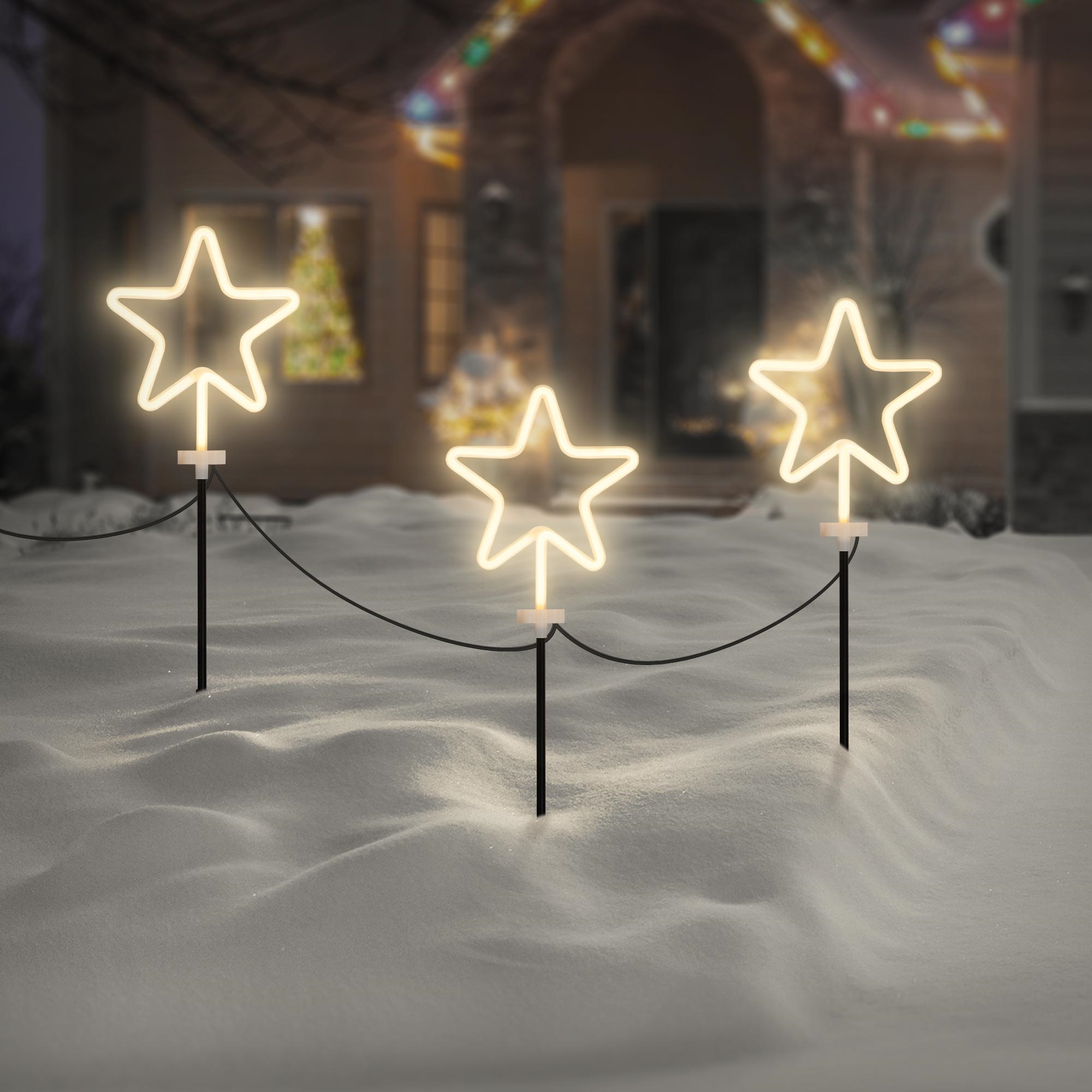Drei beleuchtete Sternverzierungen auf schwarzen Stangen im Schnee. Im Hintergrund ein Haus mit Weihnachtsbeleuchtung.