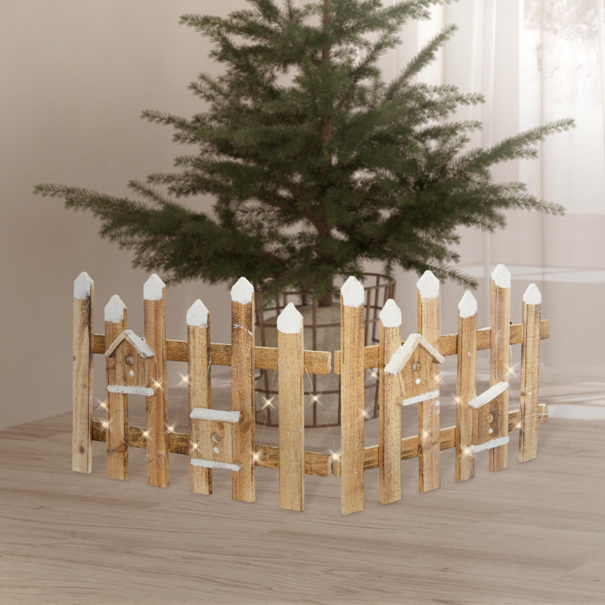 Ein kleiner Holzzaun mit Vogelhäusern umgibt einen Weihnachtsbaum. Lichter sind am Zaun aufgereiht.