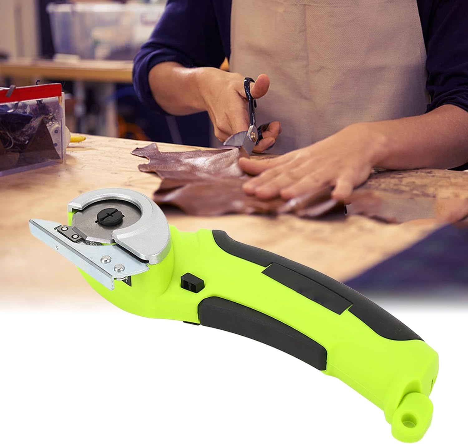 Eine Person schneidet Leder mit einer Schere und einem grünen Rollschneider auf einem Holztisch. Eine beige Schürze wird getragen.