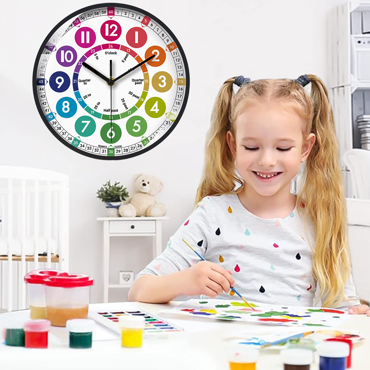 Ein Mädchen malt an einem Tisch. Eine Uhr hängt an der Wand. Farben und Pinsel liegen auf dem Tisch. Ein Bär steht auf dem kleinen Tisch.