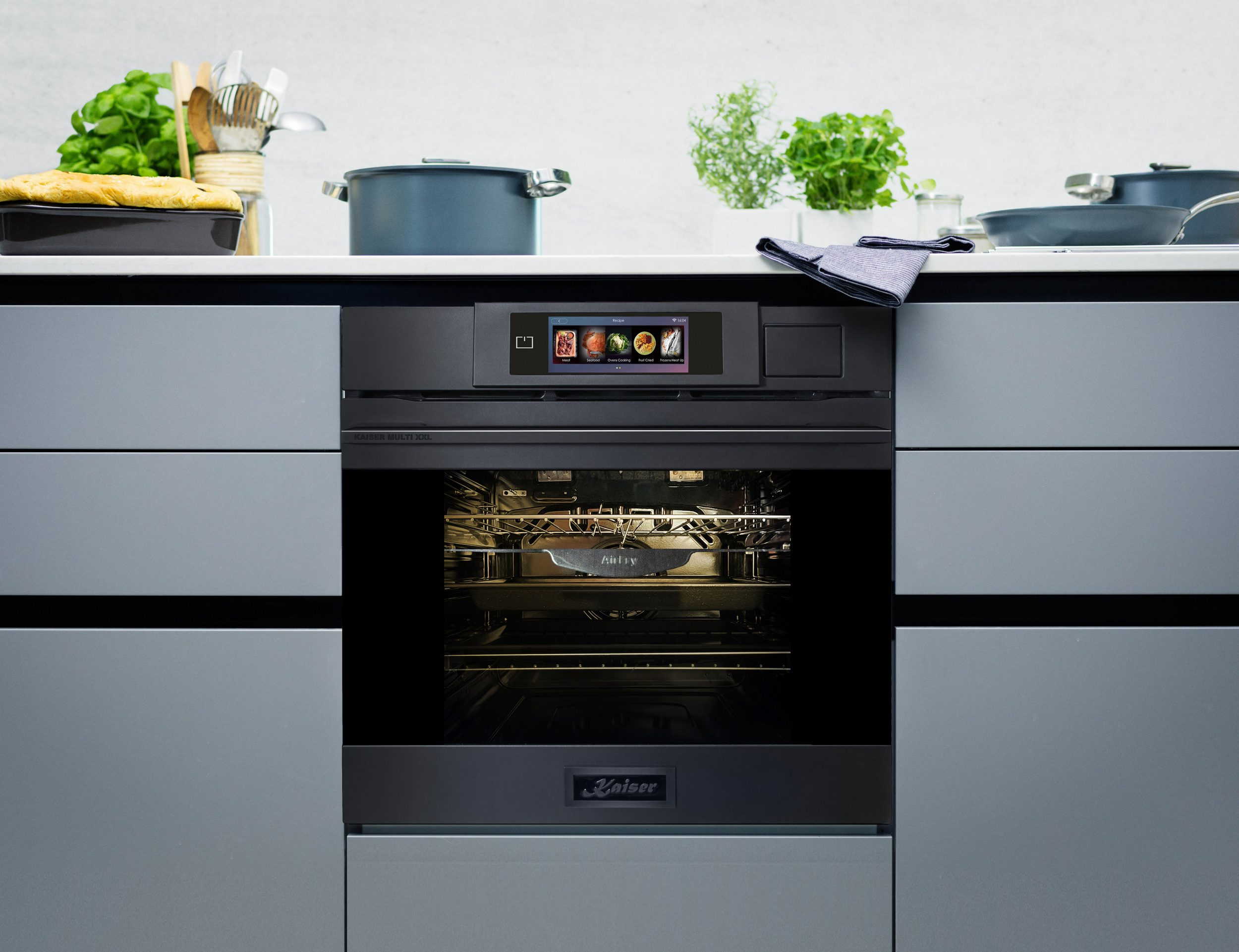 Ein moderner schwarzer Backofen ist in einen Küchenschrank eingebaut, mit Essen darin. Töpfe, Kräuter und Kochutensilien sind oben.