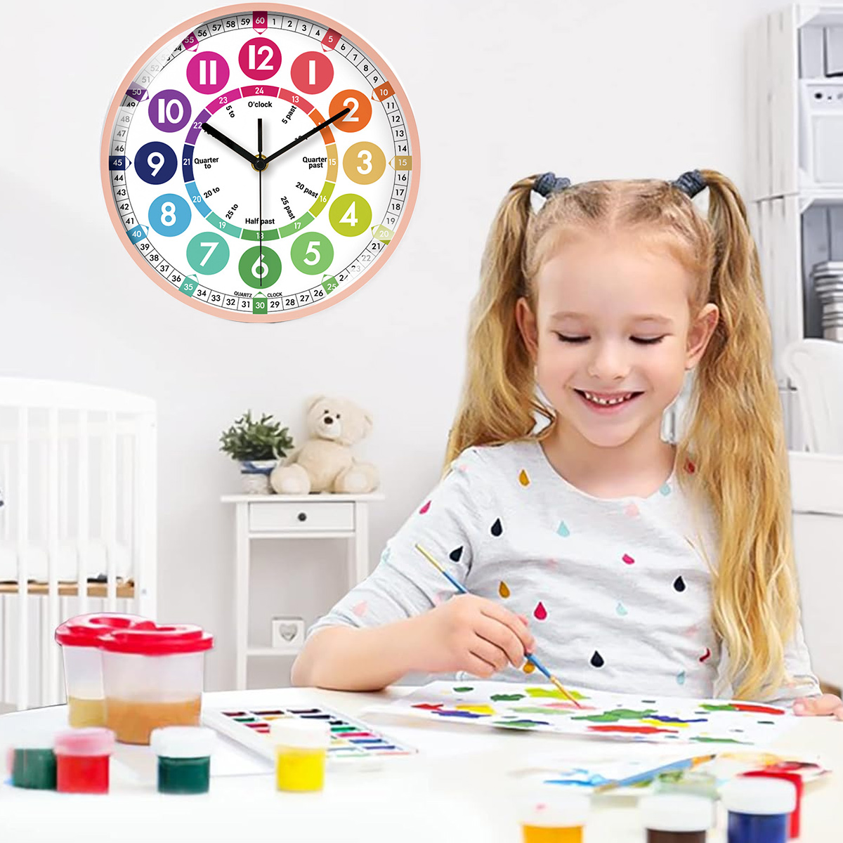 Ein Mädchen malt an einem Tisch, eine Uhr hängt an der Wand und Malutensilien sind in der Nähe. Ein Teddybär sitzt auf einem Beistelltisch.