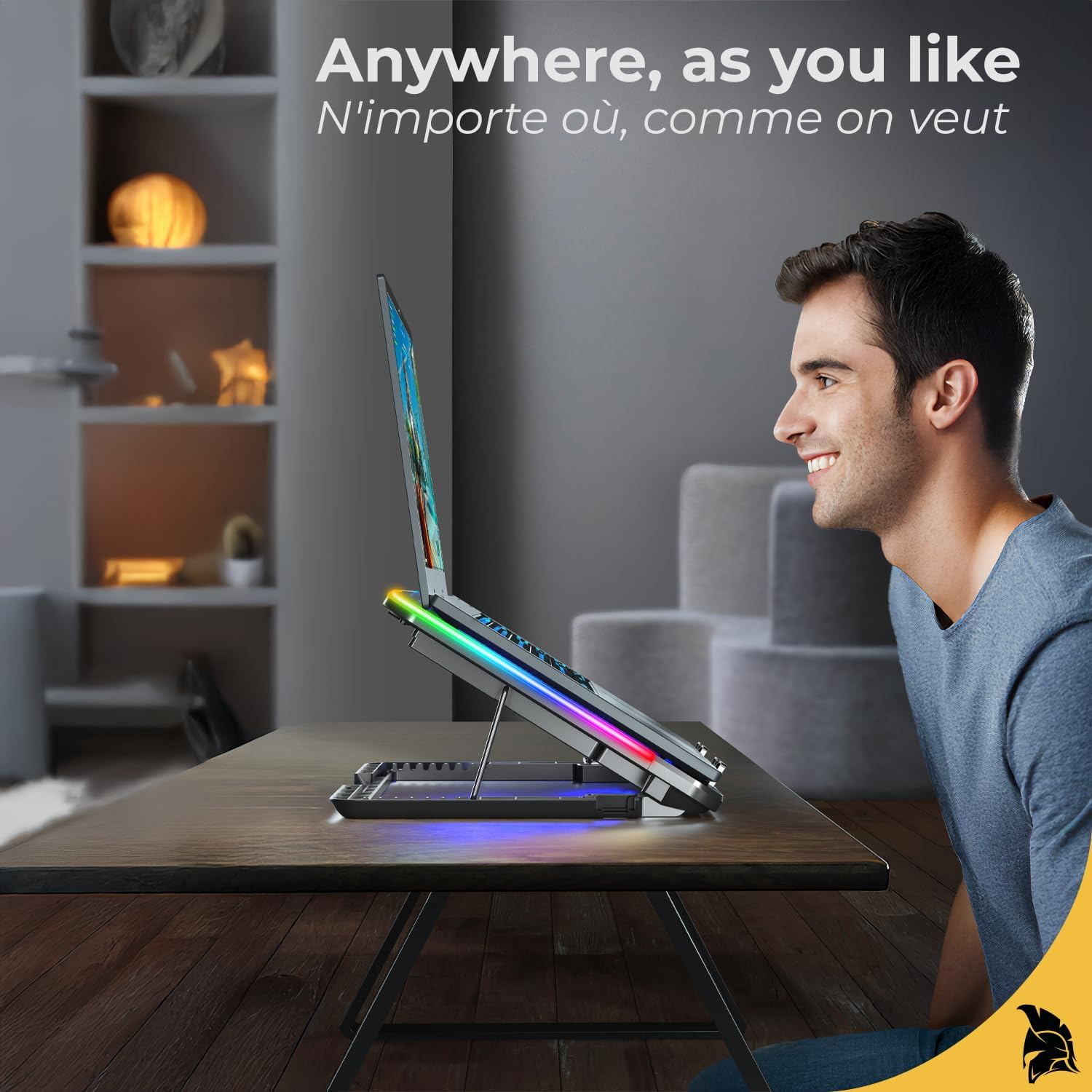 Ein Mann lächelt einen Laptop auf einem Ständer mit bunten Lichtern an. Der Laptop steht auf einem Holztisch, im Hintergrund ist ein Raum zu sehen.
