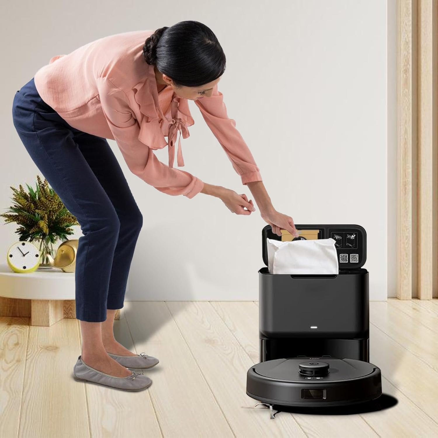 Eine Frau in rosa Oberteil und blauer Hose legt einen weißen Beutel in eine schwarze Roboter-Staubsaugerbasis.