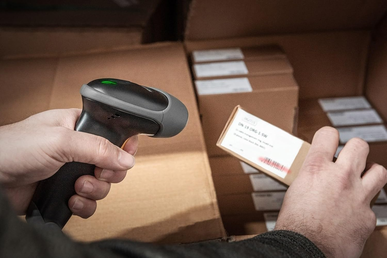 Eine Person scannt ein Etikett auf einer Schachtel mit einem Barcode-Scanner. Im Hintergrund befinden sich weitere Kartons.