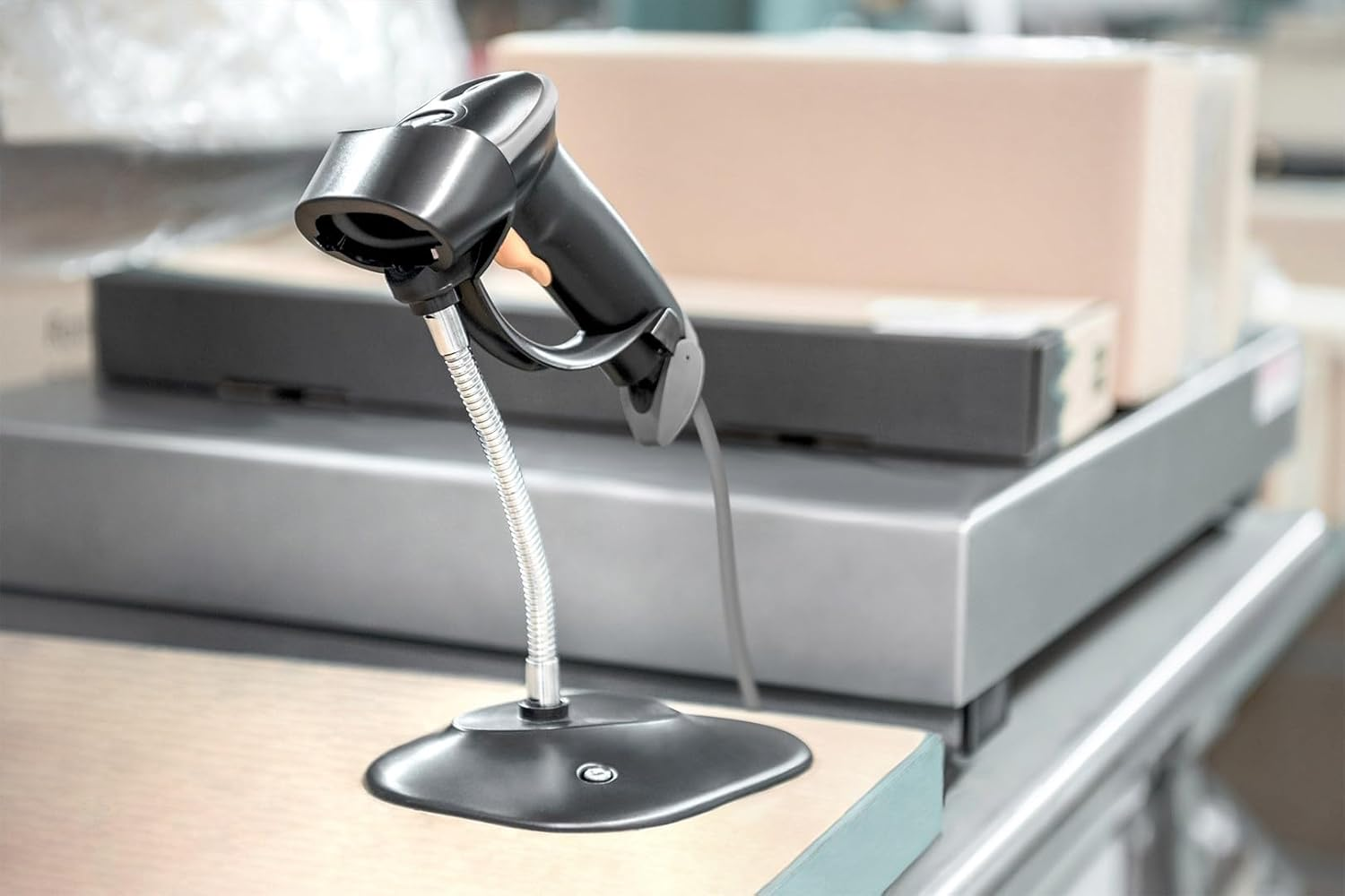 Ein schwarzer Barcode-Scanner auf einem Ständer, Kisten im Hintergrund. Der Scanner steht auf einem Tisch. Die Szene spielt in einem Lager.