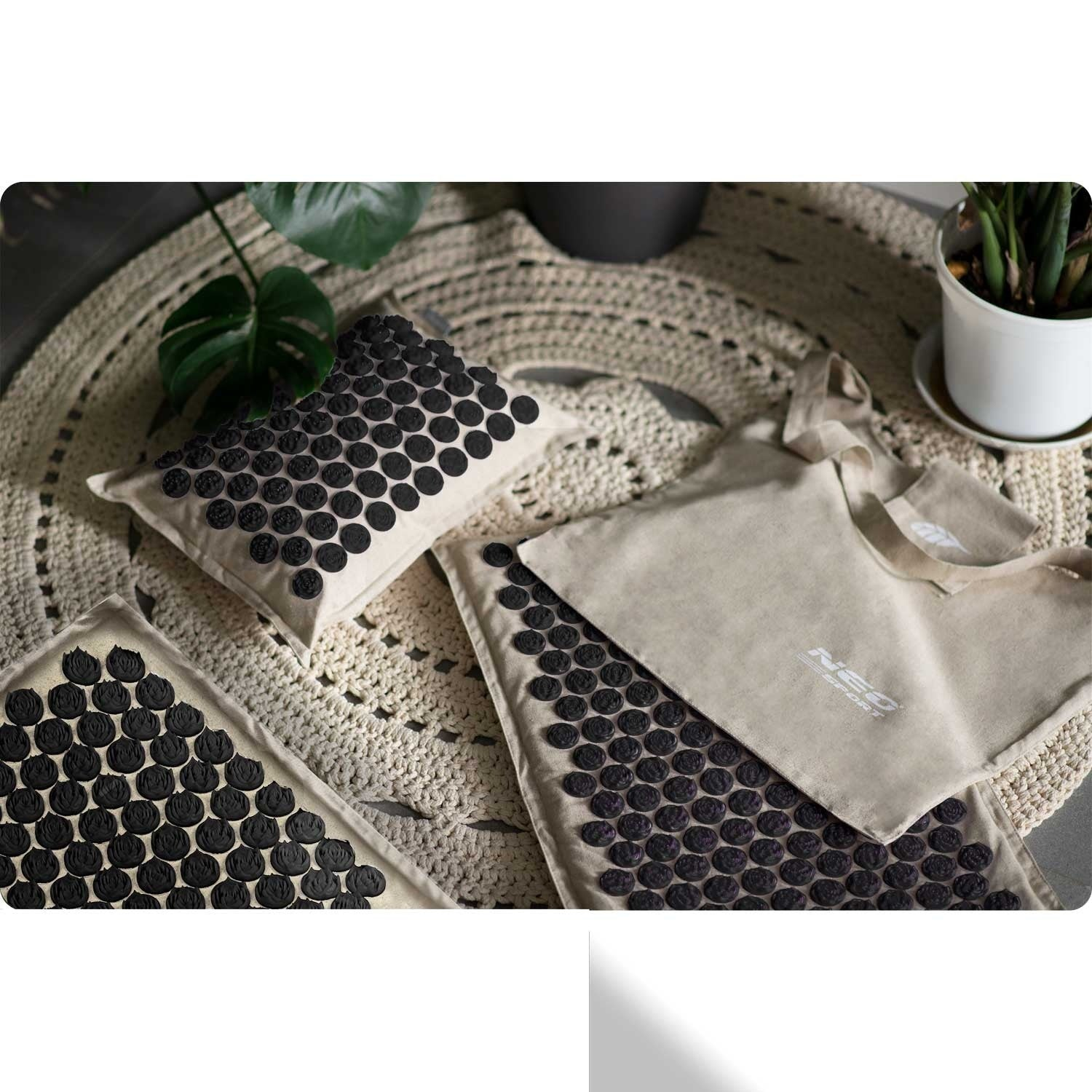 Una esterilla beige con elementos de masaje con púas negras, una almohada, una bolsa y plantas sobre una alfombra beige.