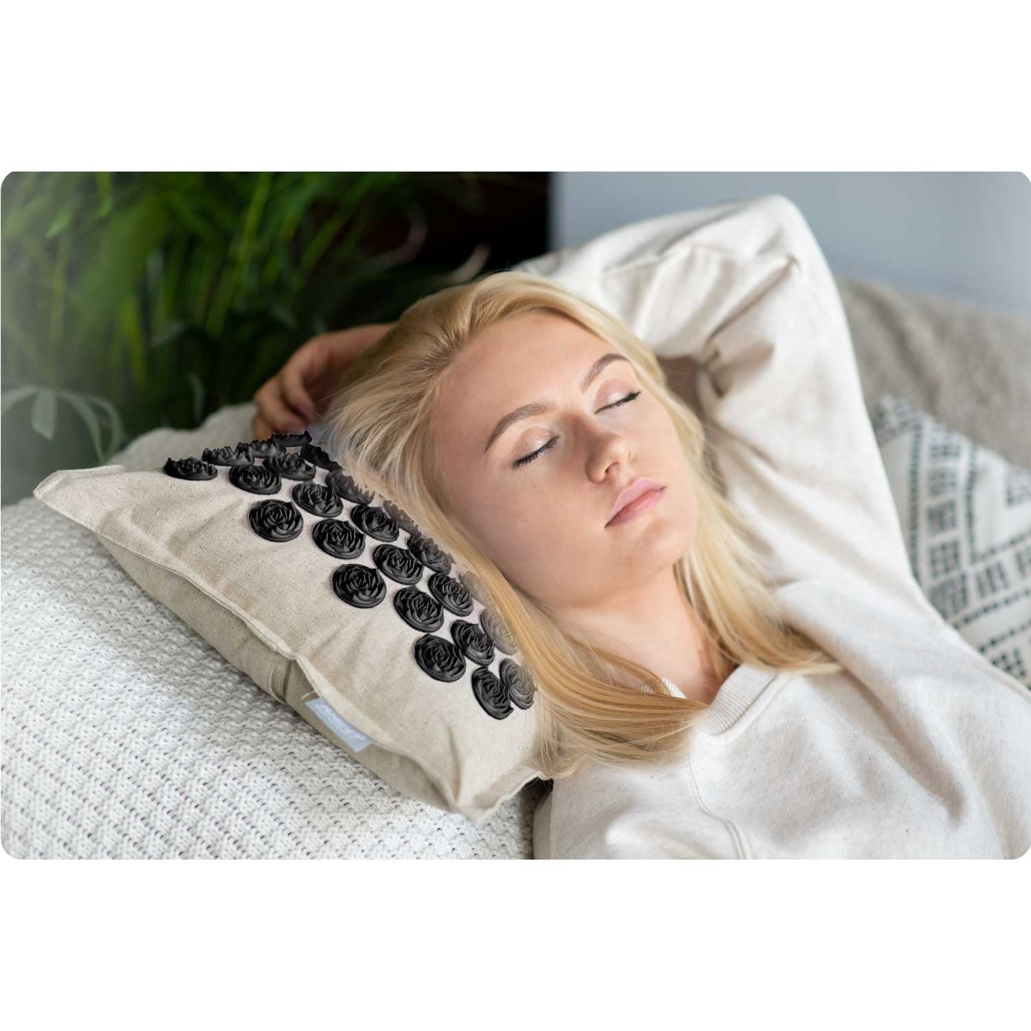 Una mujer rubia descansa en un sofá con una almohada de masaje. Tiene los ojos cerrados. Hay una planta en el fondo.