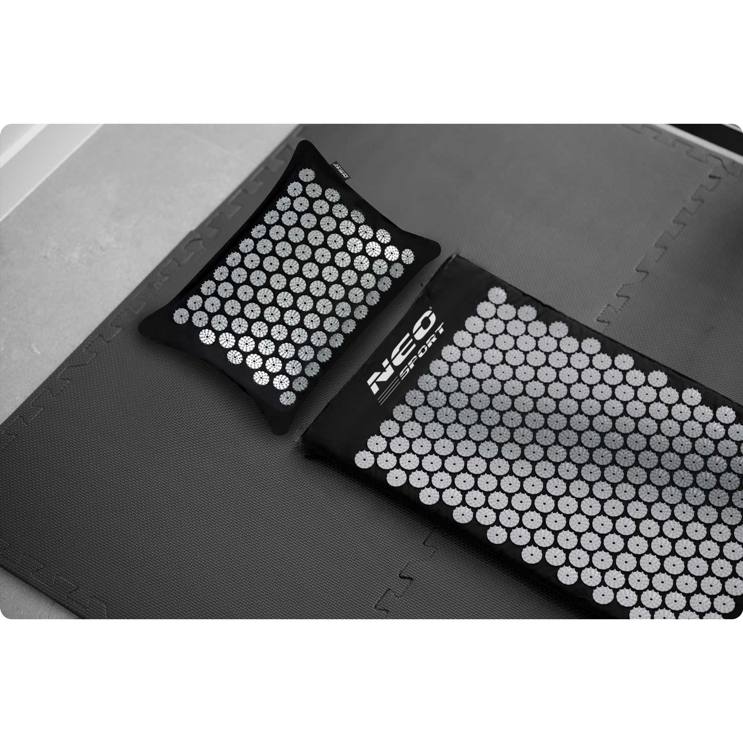 Dos esterillas de masaje negras con puntas blancas sobre una esterilla gris. Una es una almohada, la otra una esterilla rectangular más grande, ambas con el logotipo de Neo Sport.