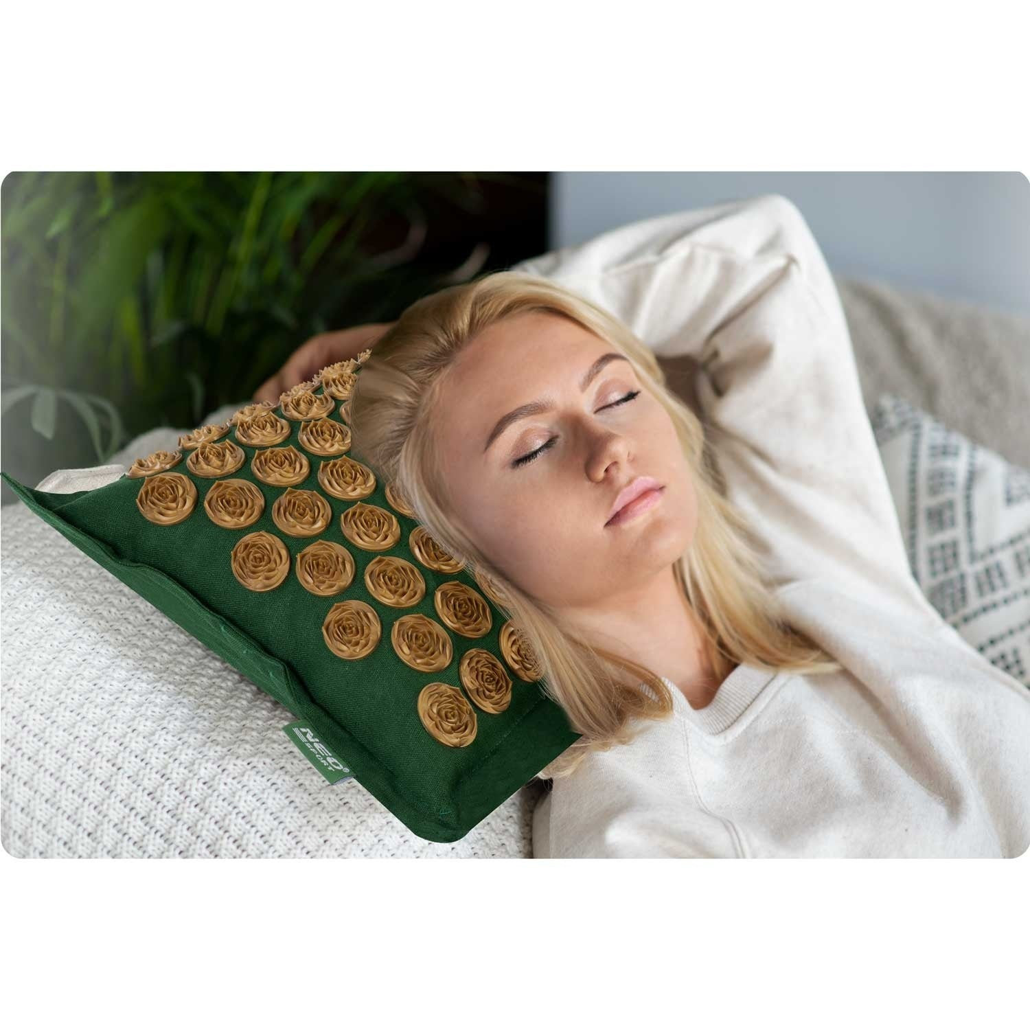 Una mujer rubia yace en un sofá, apoyando su cabeza en una almohada verde con adornos dorados en forma de rosa.