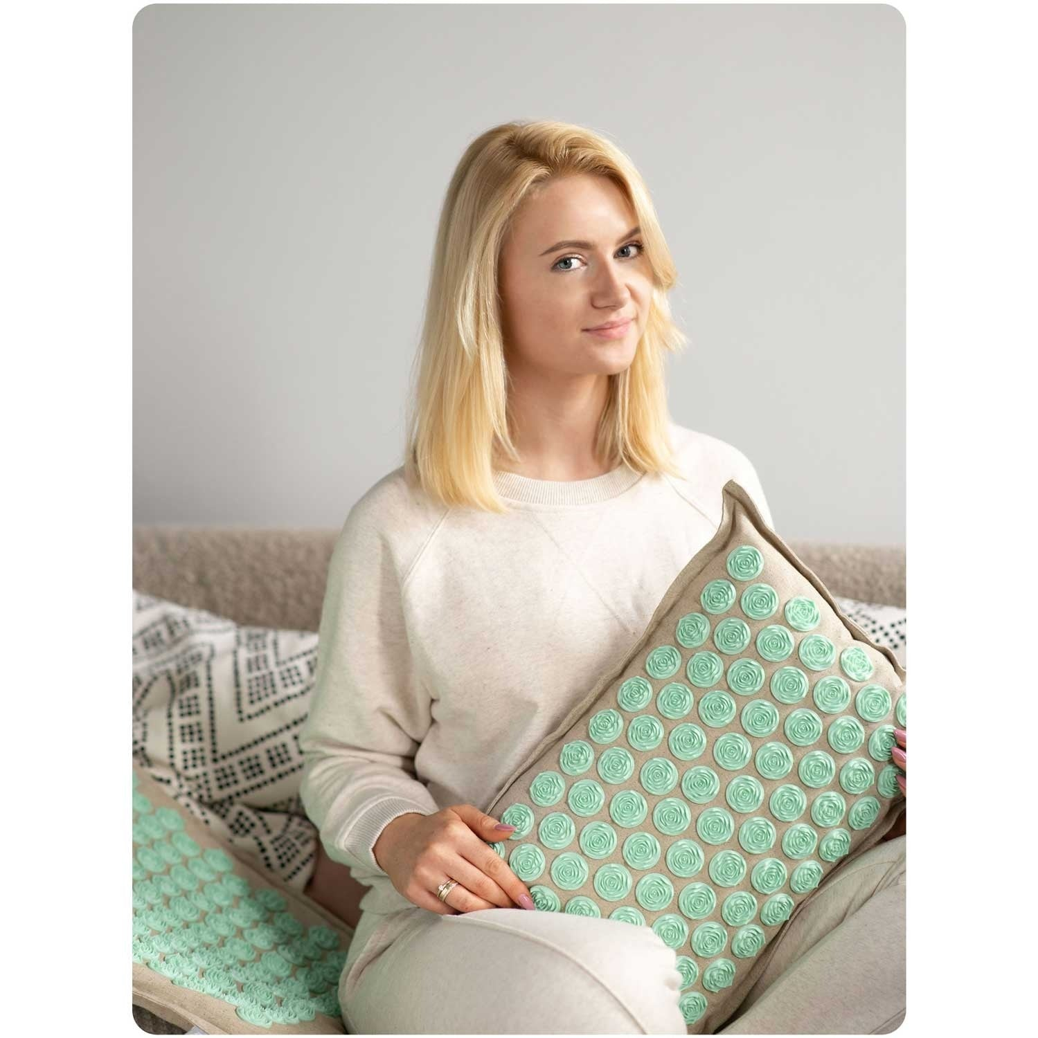 Mujer rubia sosteniendo una almohada de masaje verde menta con puntos en forma de rosa, sentada en un sofá. Sonríe a la cámara.