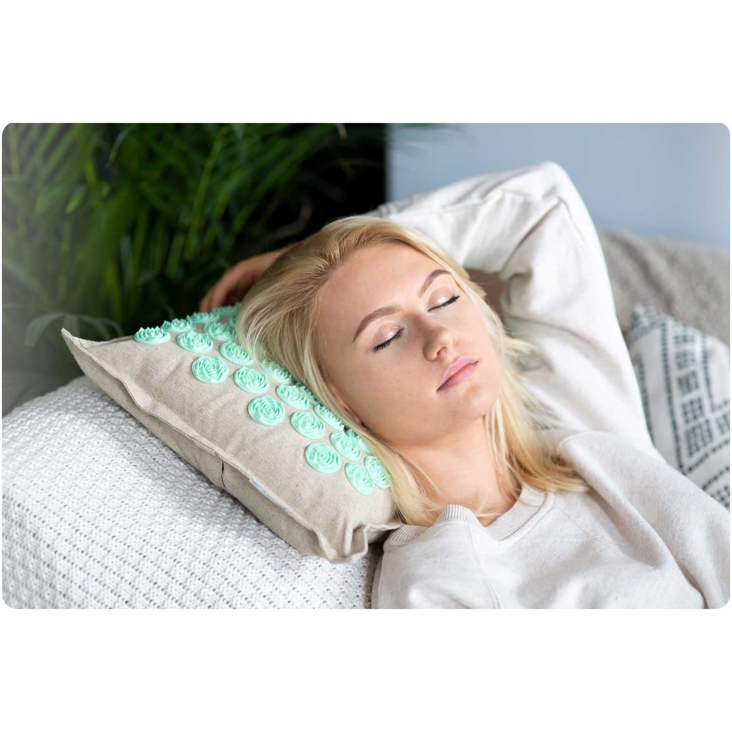Una mujer con cabello rubio yace en un sofá, apoyando la cabeza en una almohada con puntos verdes de acupresión.