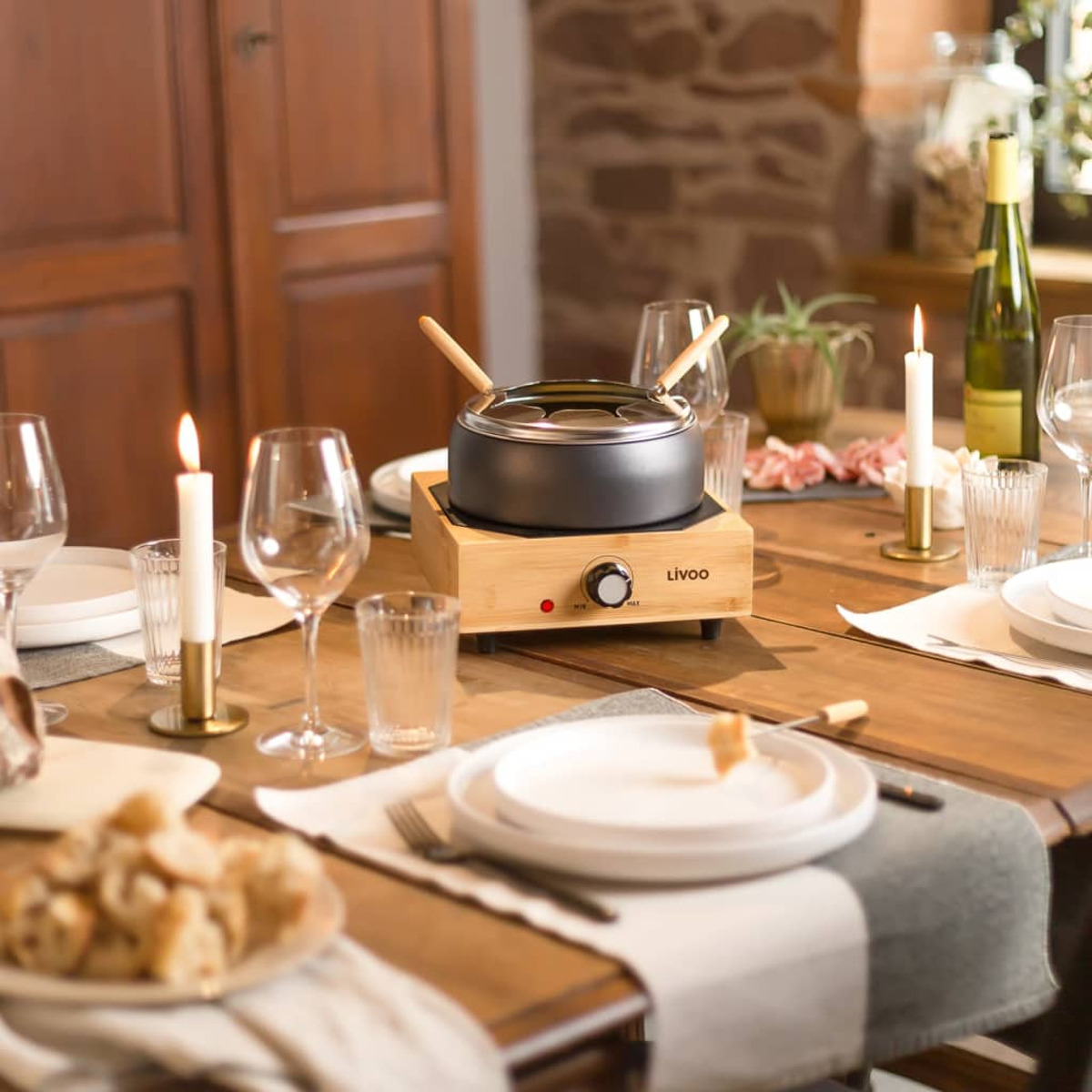 Ein Tisch ist für Fondue gedeckt, mit Topf, Kerzen, Gläsern, Tellern und Essen. Im Hintergrund steht eine Weinflasche.