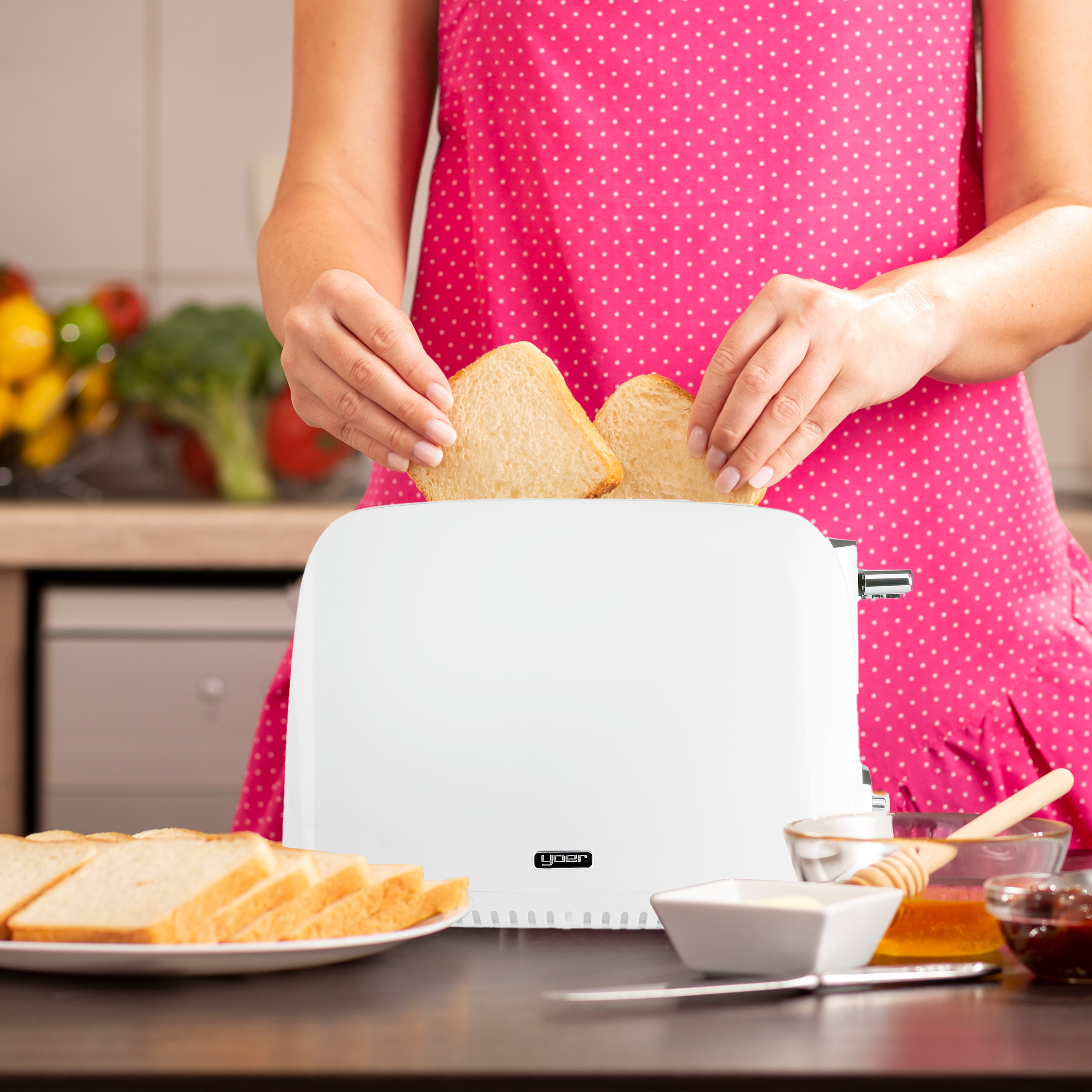 Osoba wkłada kromki chleba do białego tostera. Na stole znajdują się kromki chleba, masło, miód i dżem.