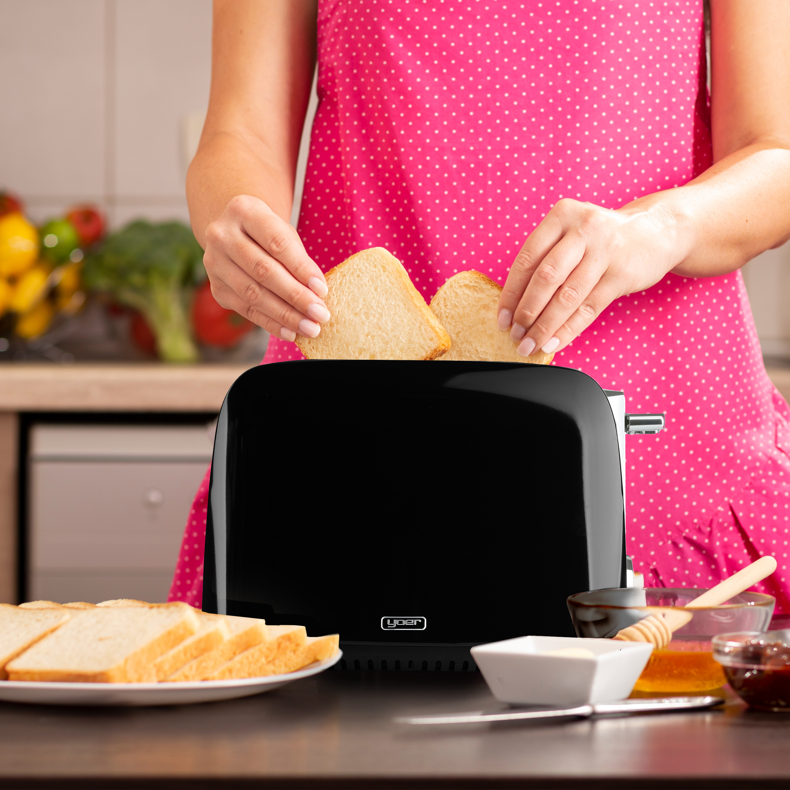 Osoba w różowym fartuchu wkłada kromki chleba do czarnego tostera. Chleb, masło, miód i dżem na stole.