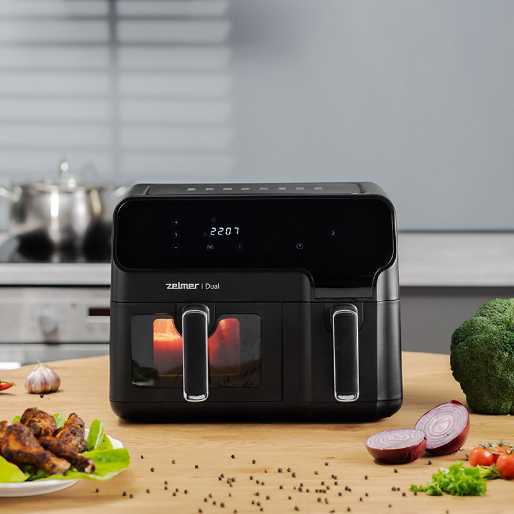 Una friggitrice ad aria nera è su un tavolo di legno con cibi come pollo, verdure e spezie. Lo sfondo mostra una cucina.