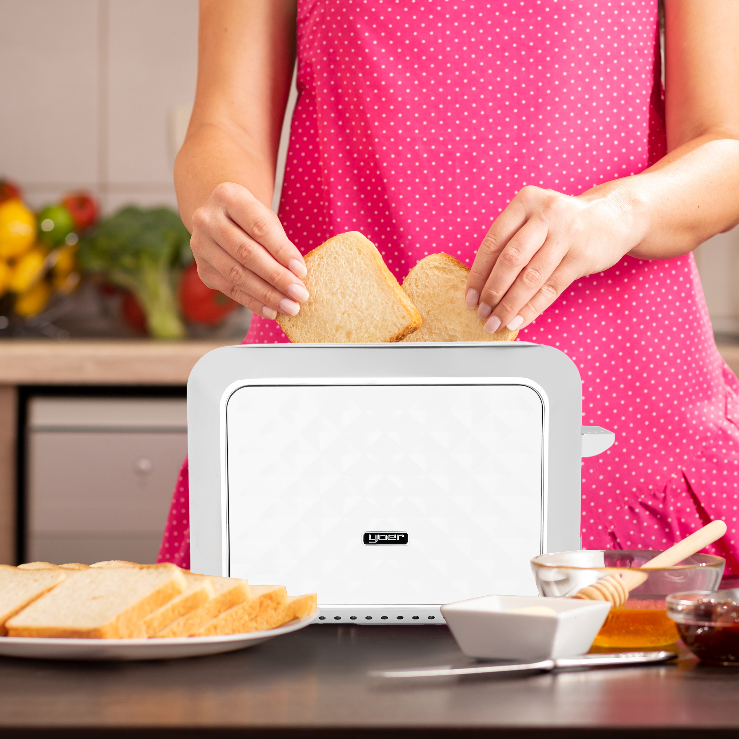Osoba w różowym fartuchu w grochy wkłada chleb do białego tostera. Na stole kromki chleba, masło, miód i dżem.